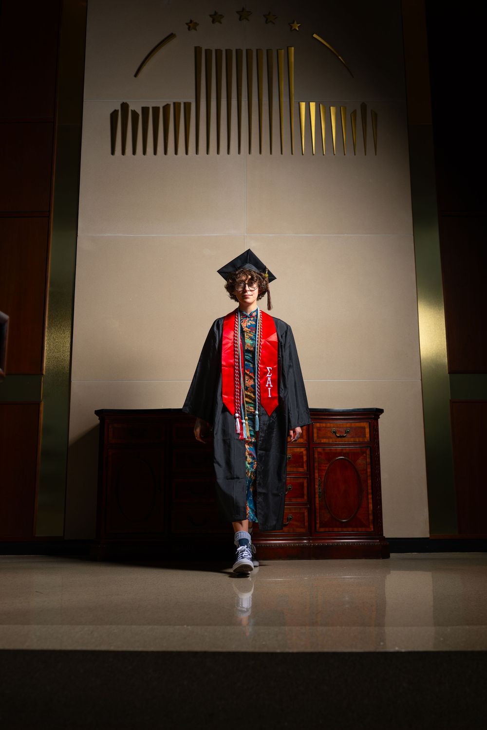 Graduate in black cap and gown with red stole walks across stage at indoor graduation ceremony.