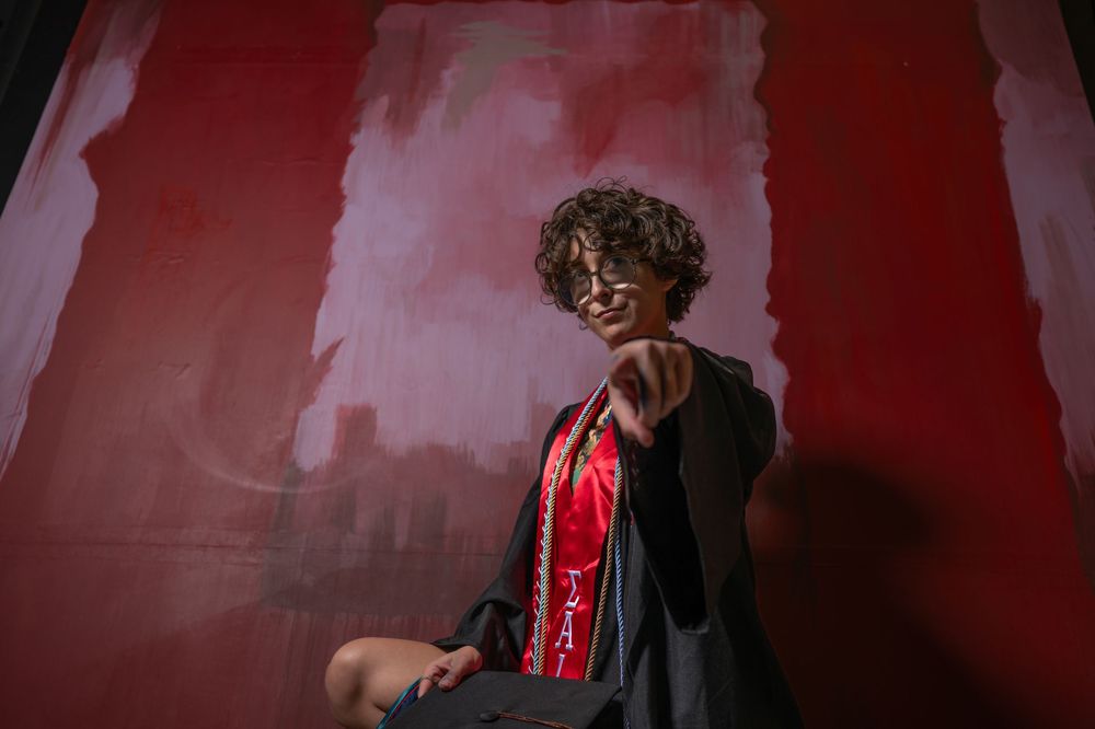 Graduate in black gown and red sash poses confidently against a red painted backdrop during graduation photo session.