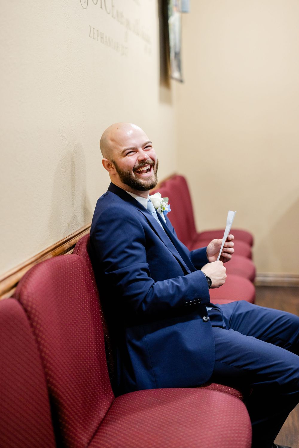 A person in a navy suit sits on a red upholstered bench while looking at their phone with a joyful expression.