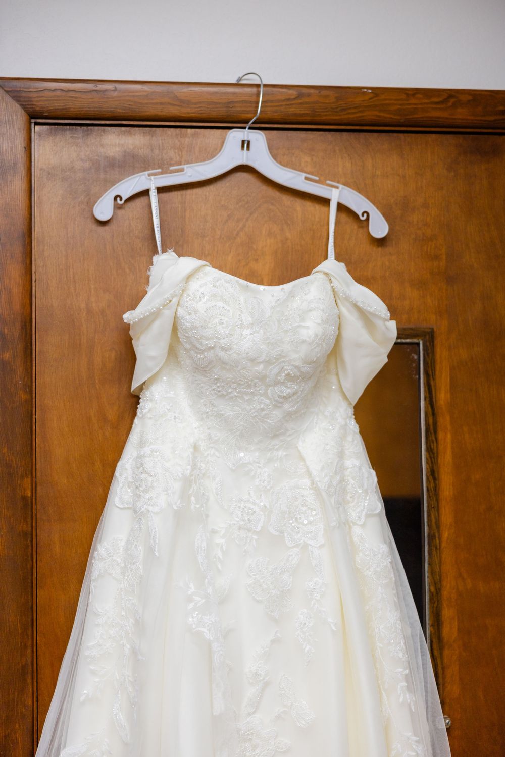 A white lace wedding dress with off-shoulder sleeves hangs on a white plastic hanger against a wooden door.