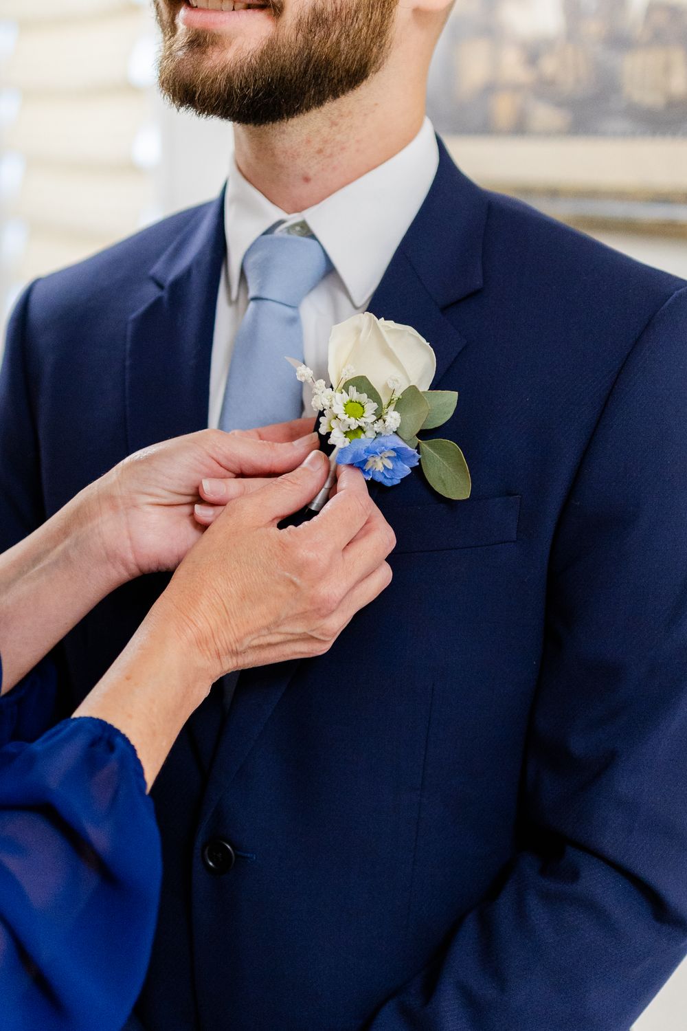 Hands pinning a delicate blue and white floral boutonniere onto a navy blue suit jacket.
