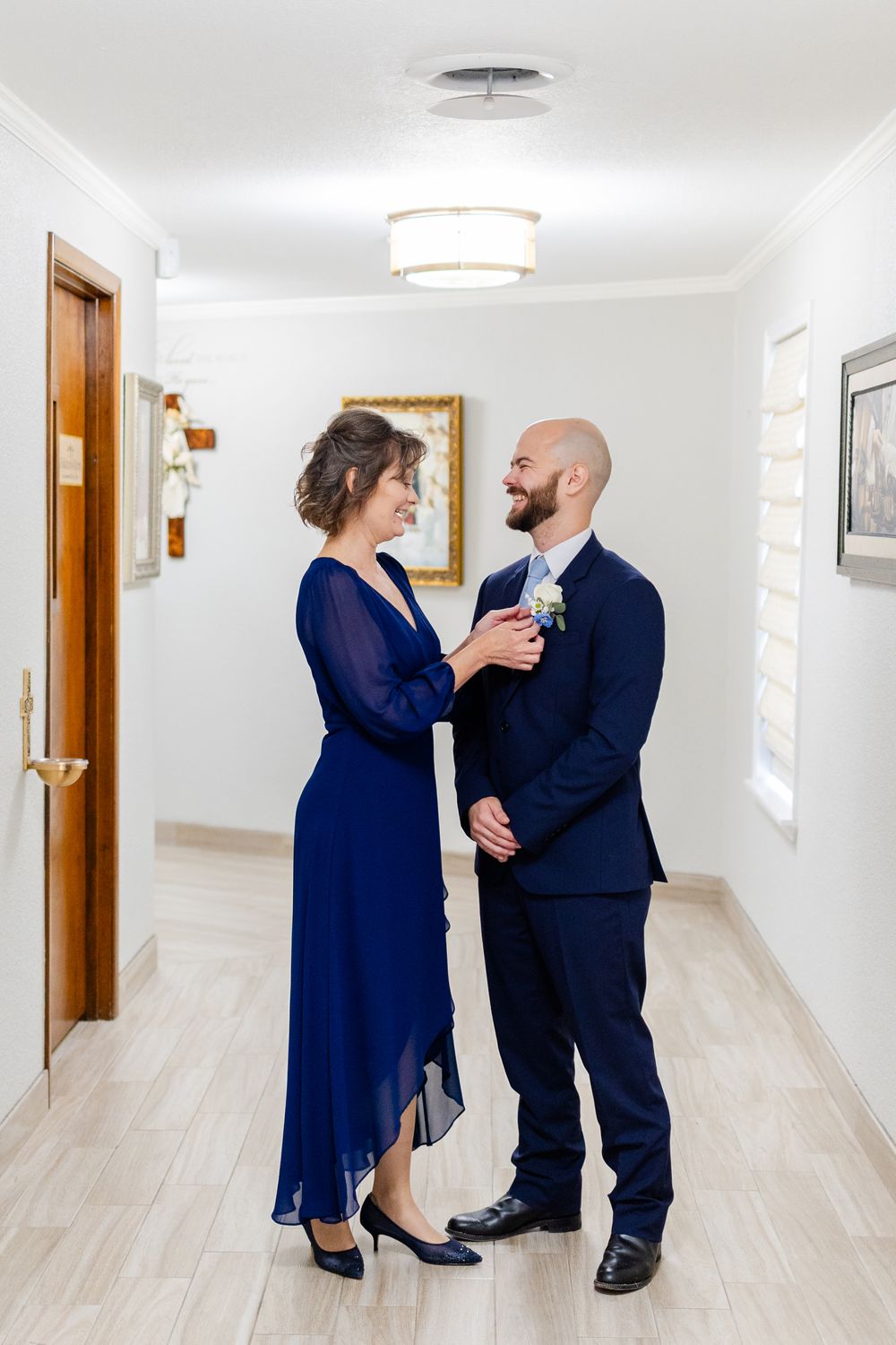 Two people in elegant navy blue formal wear stand together in a bright hallway with light wood floors.