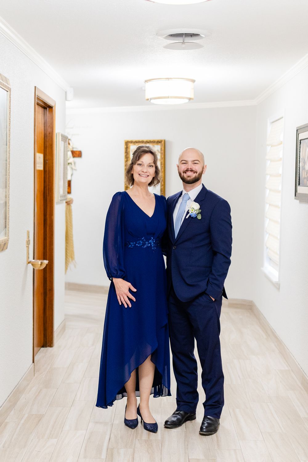 A couple in formal attire poses in a bright hallway, with the woman wearing a navy dress and man in a navy suit.