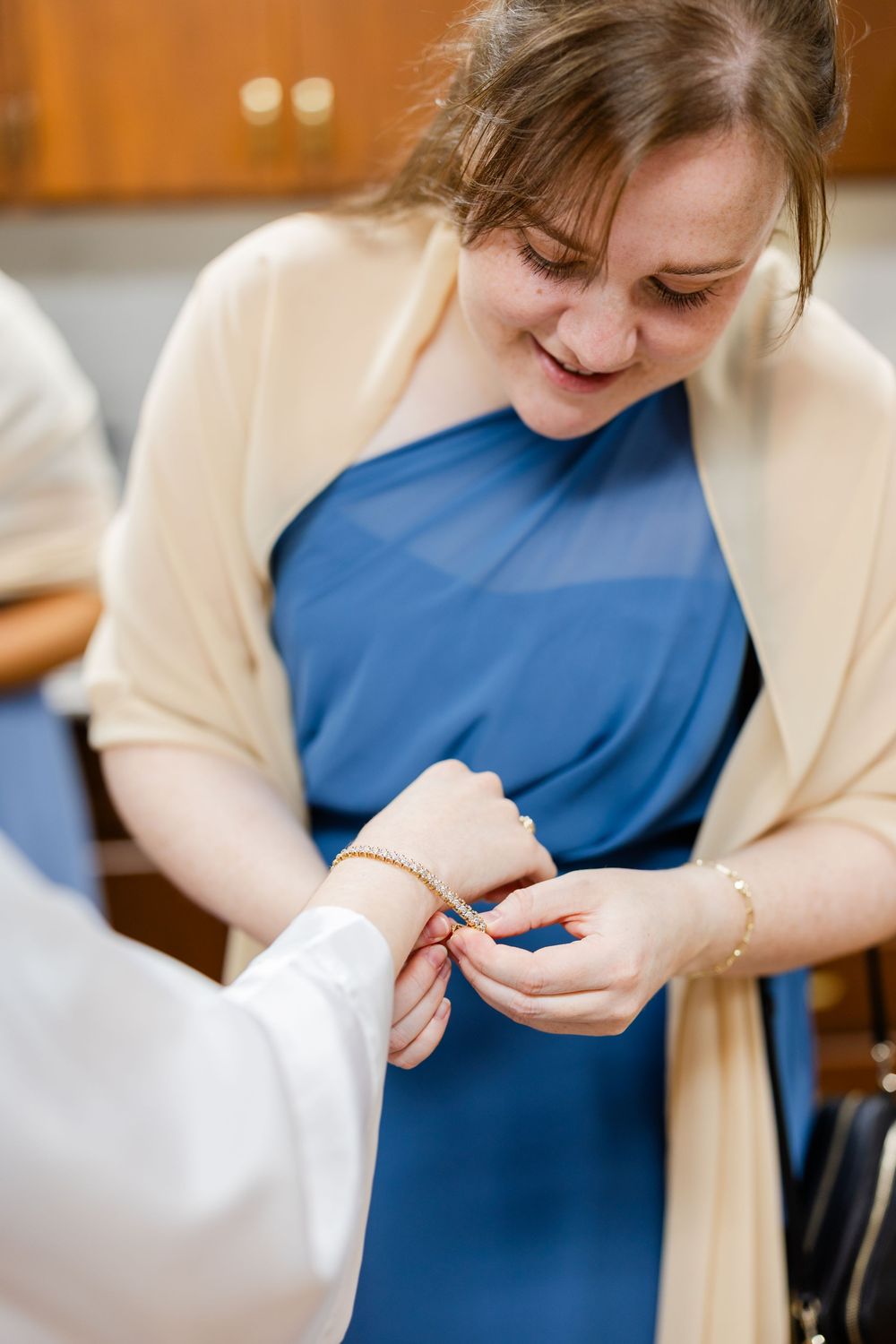 Someone in a blue dress and cream cardigan having jewelry adjusted during wedding preparations.