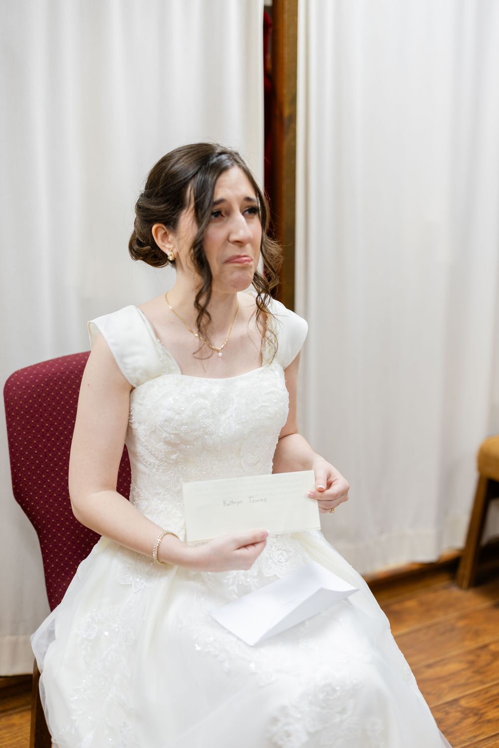 A bride in a white wedding dress sits in a red chair holding a letter before the ceremony.