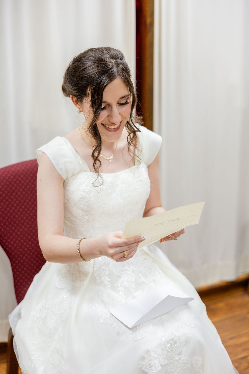 A bride in a white lace wedding gown smiles while reading a letter during wedding preparations.