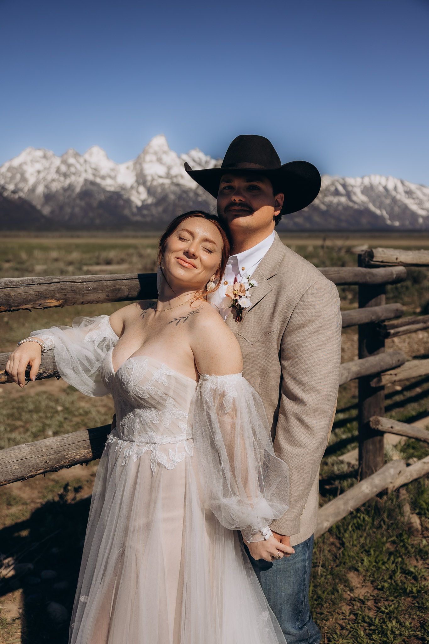 Wedding couple in western attire embracing against rustic fence with mountain backdrop.