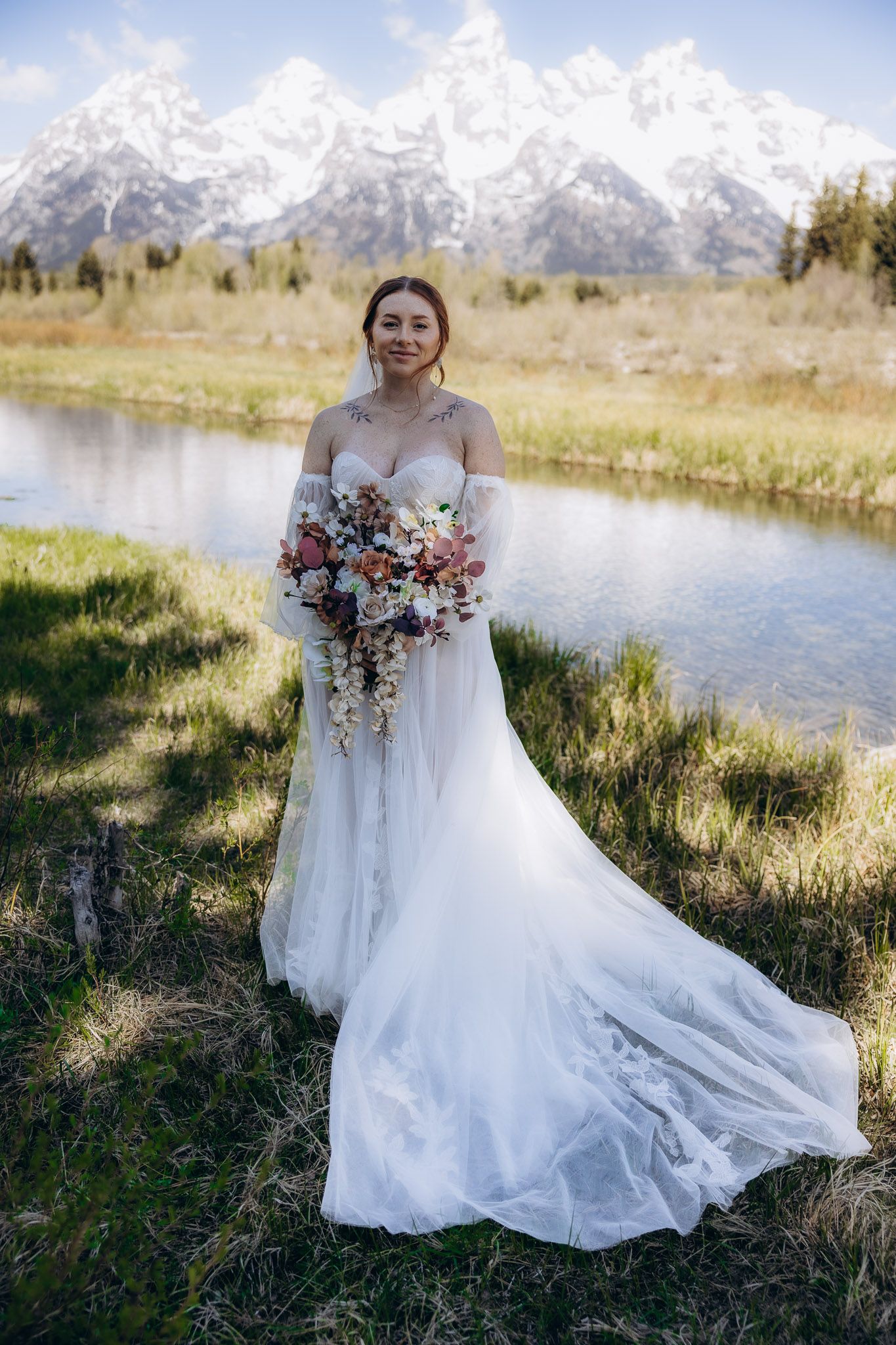 A bride in a flowing white gown holds a colorful bouquet against a backdrop of snow-capped mountains.
