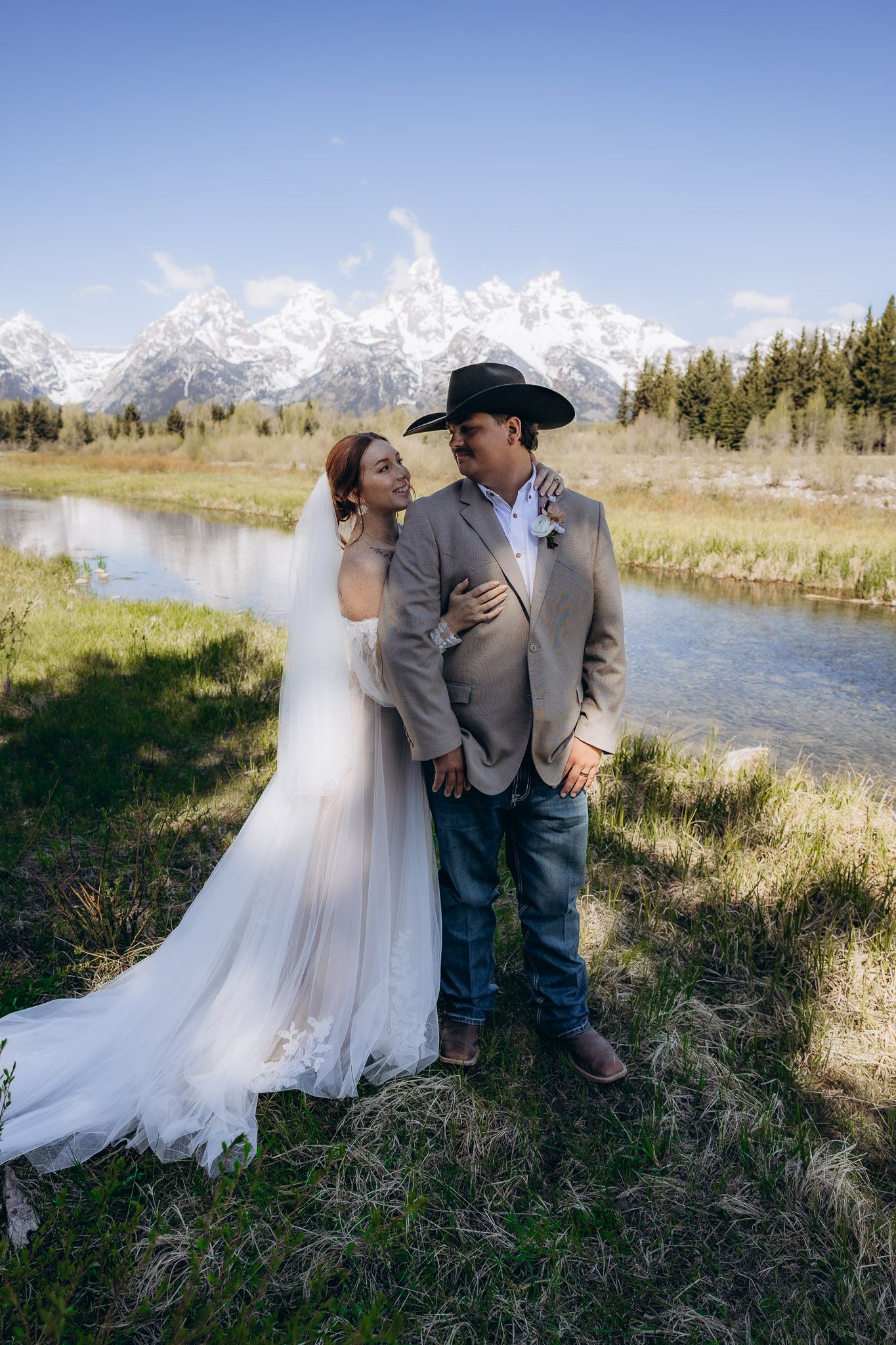 Wedding couple embraces by a river with snow-capped Grand Tetons mountains in the background.