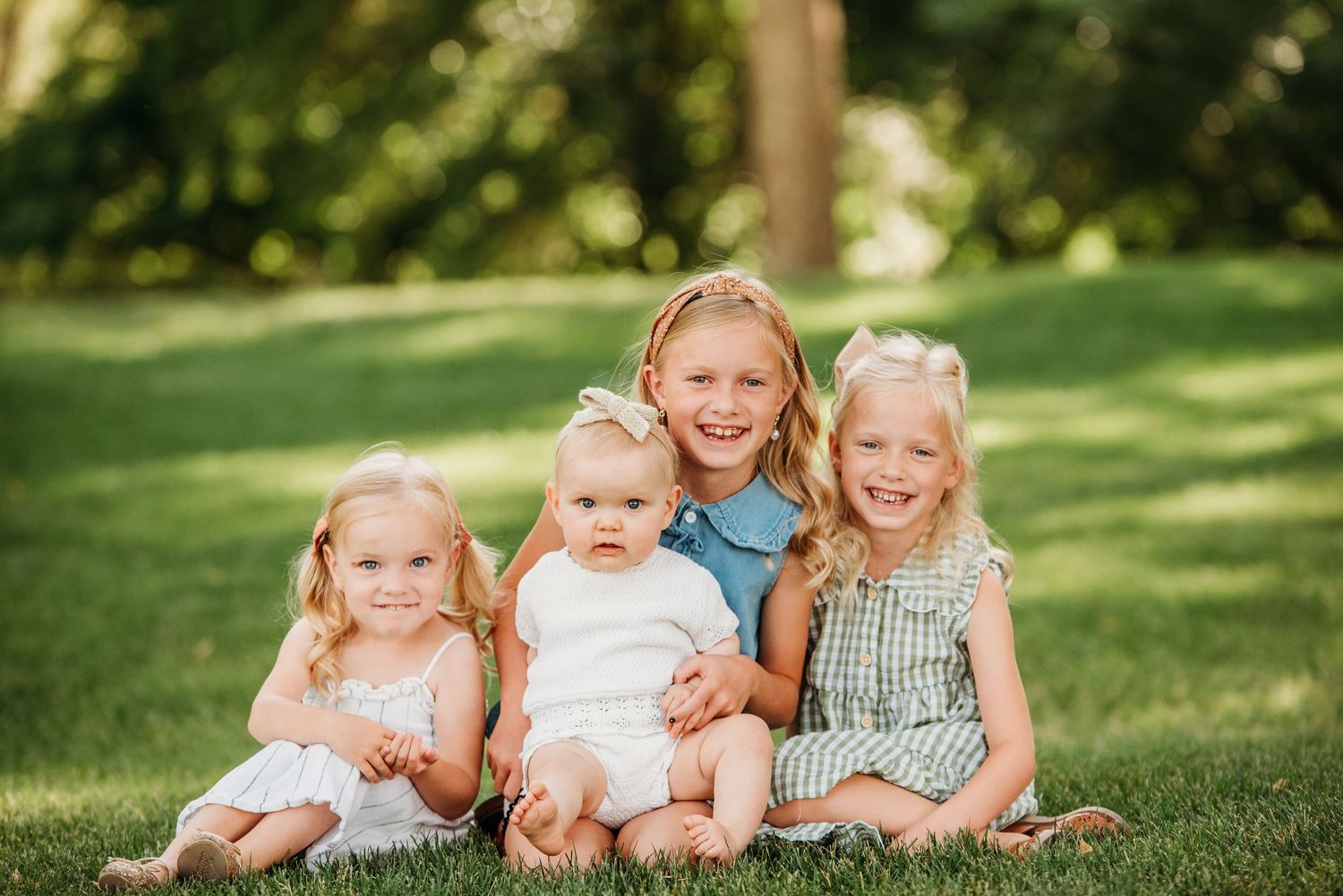 Four young children sit together on green grass in a sunlit backyard on a summer day.