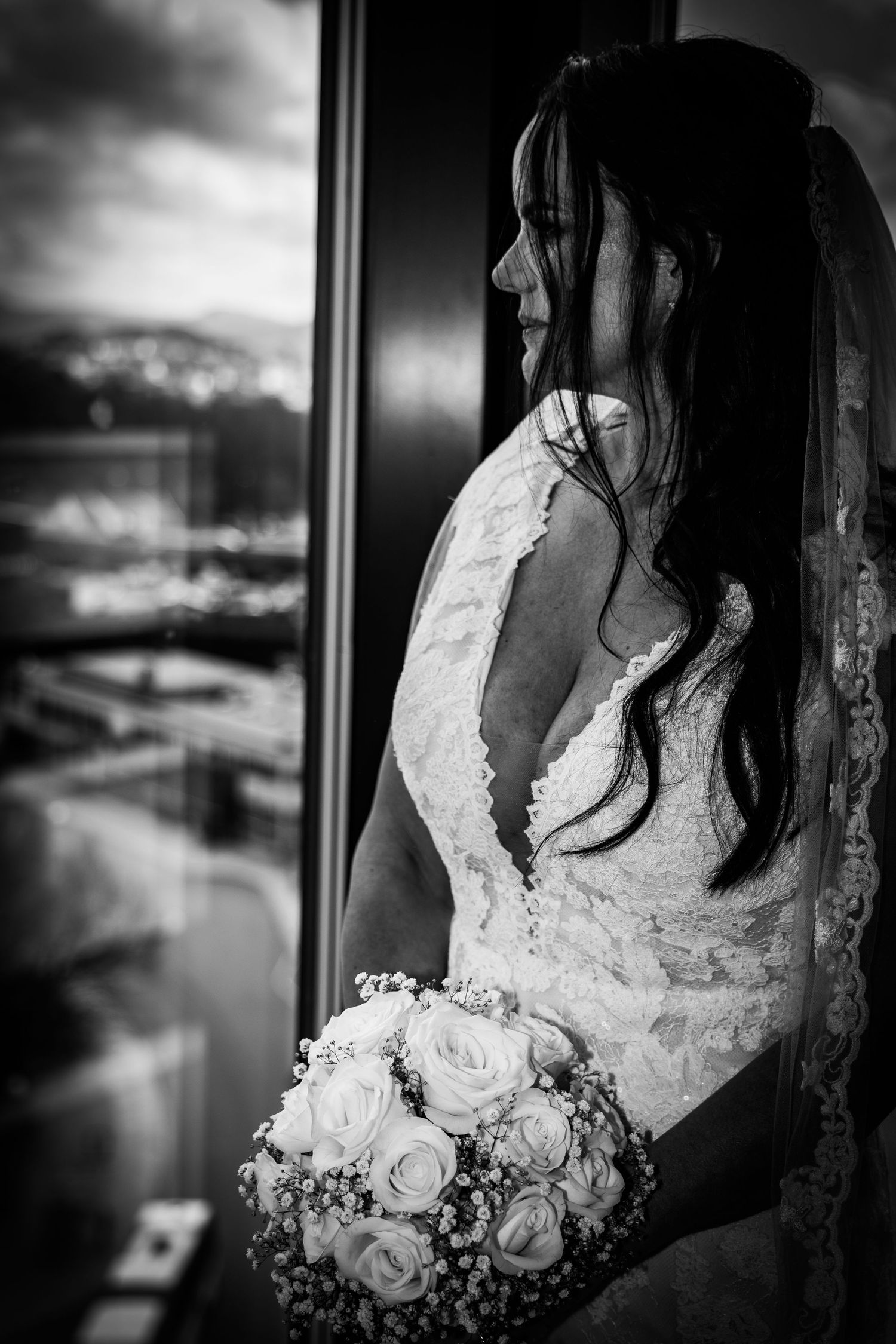 Black and white portrait of bride in lace gown holding rose bouquet gazing out hotel window.