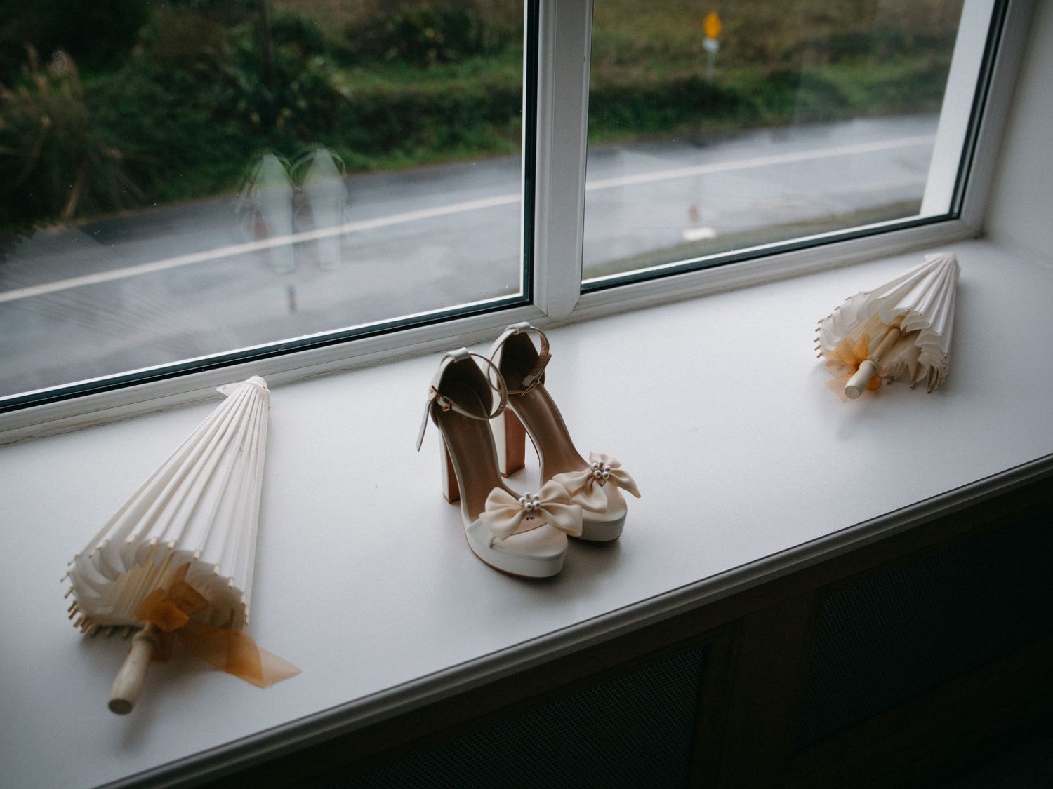 Elegant white wedding shoes sit on a window sill with decorative tassels on either side.