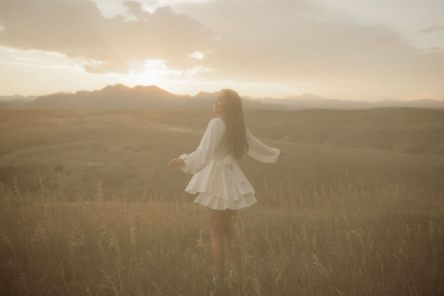 A figure in a white dress stands in a golden field at sunset, silhouetted against a dreamy mountainous landscape.