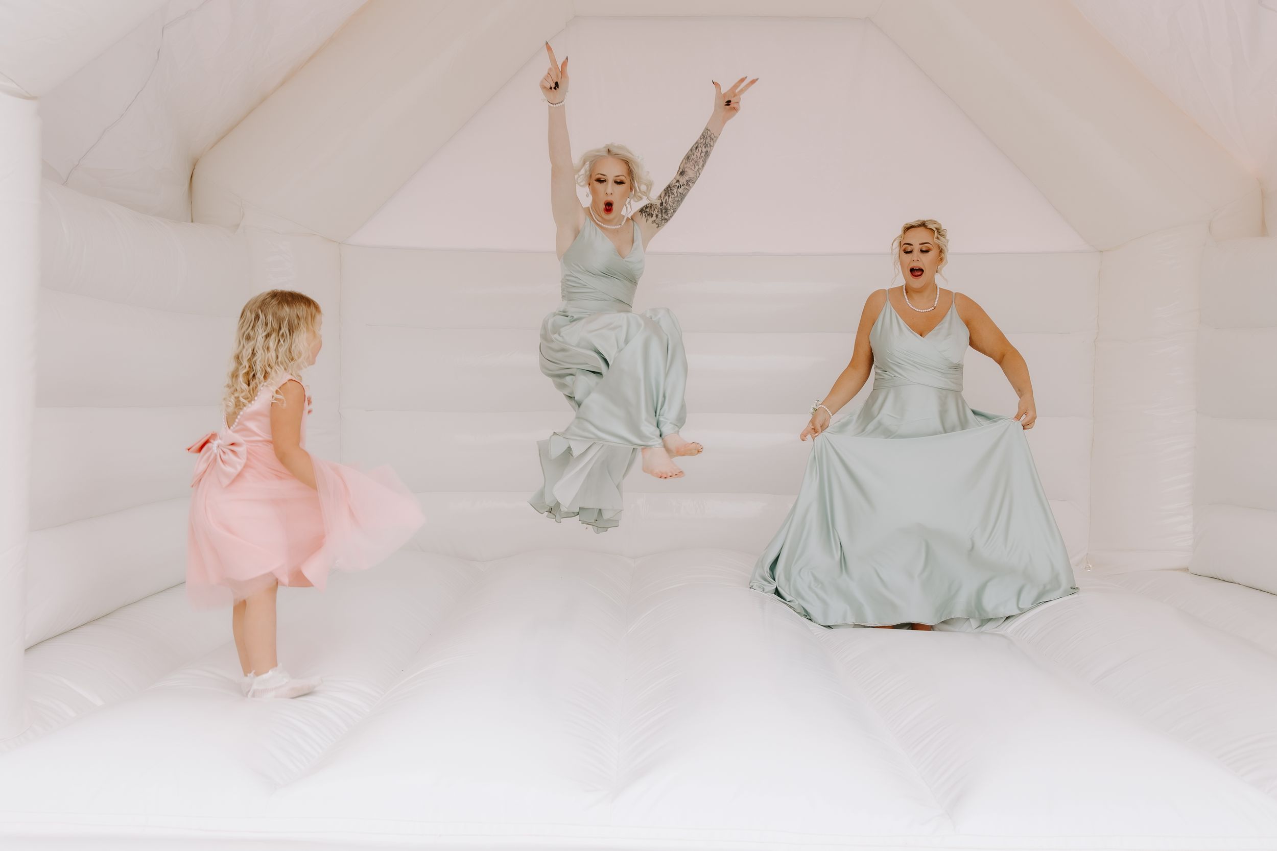 Three people in elegant pastel dresses dancing and jumping joyfully on a white bounce house surface.