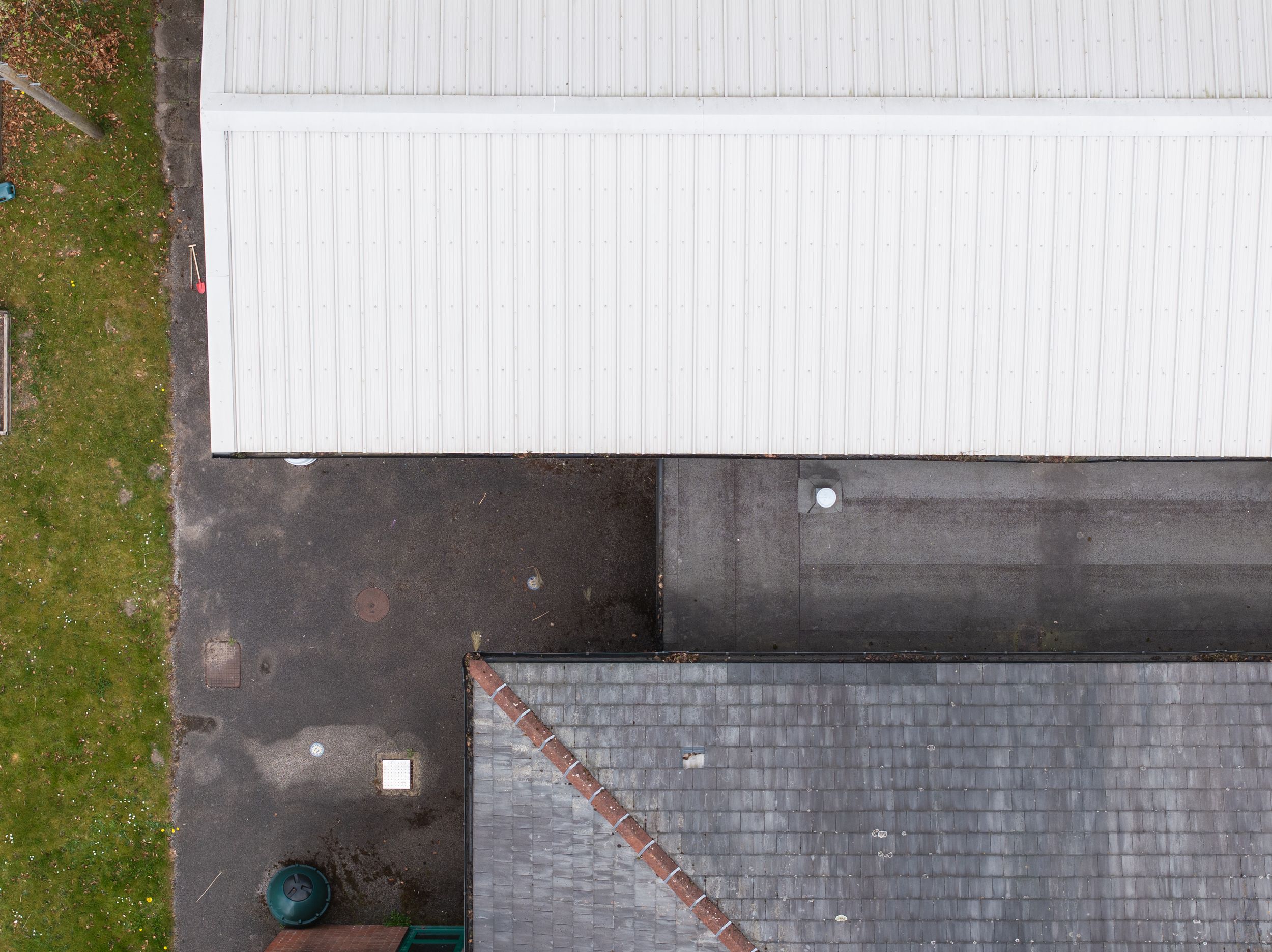 Aerial view showing corner of white building roof meeting dark shingle roofing with grass edge visible.