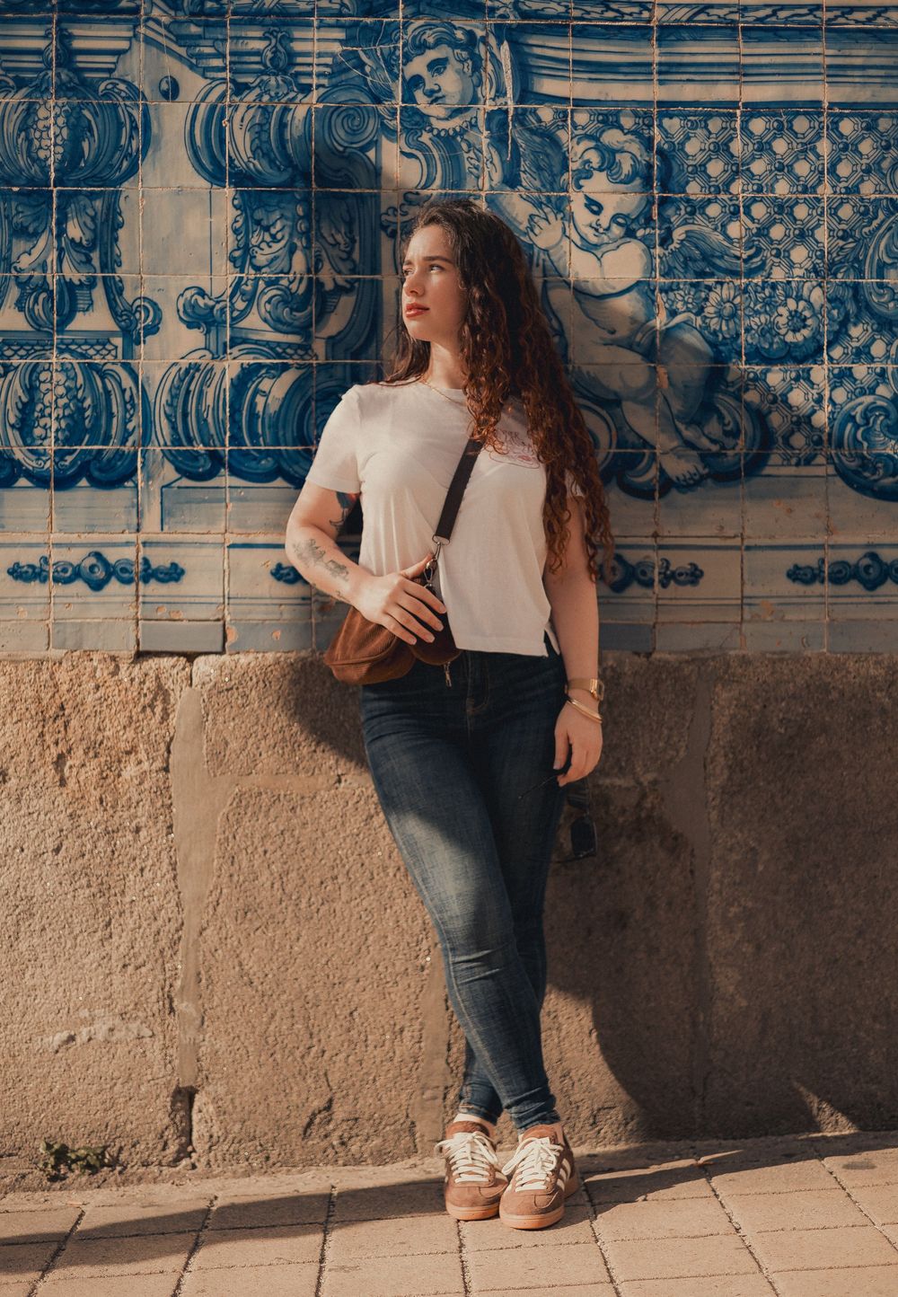 Portrait against blue Portuguese azulejo tiles wearing white top and jeans with long curly hair.