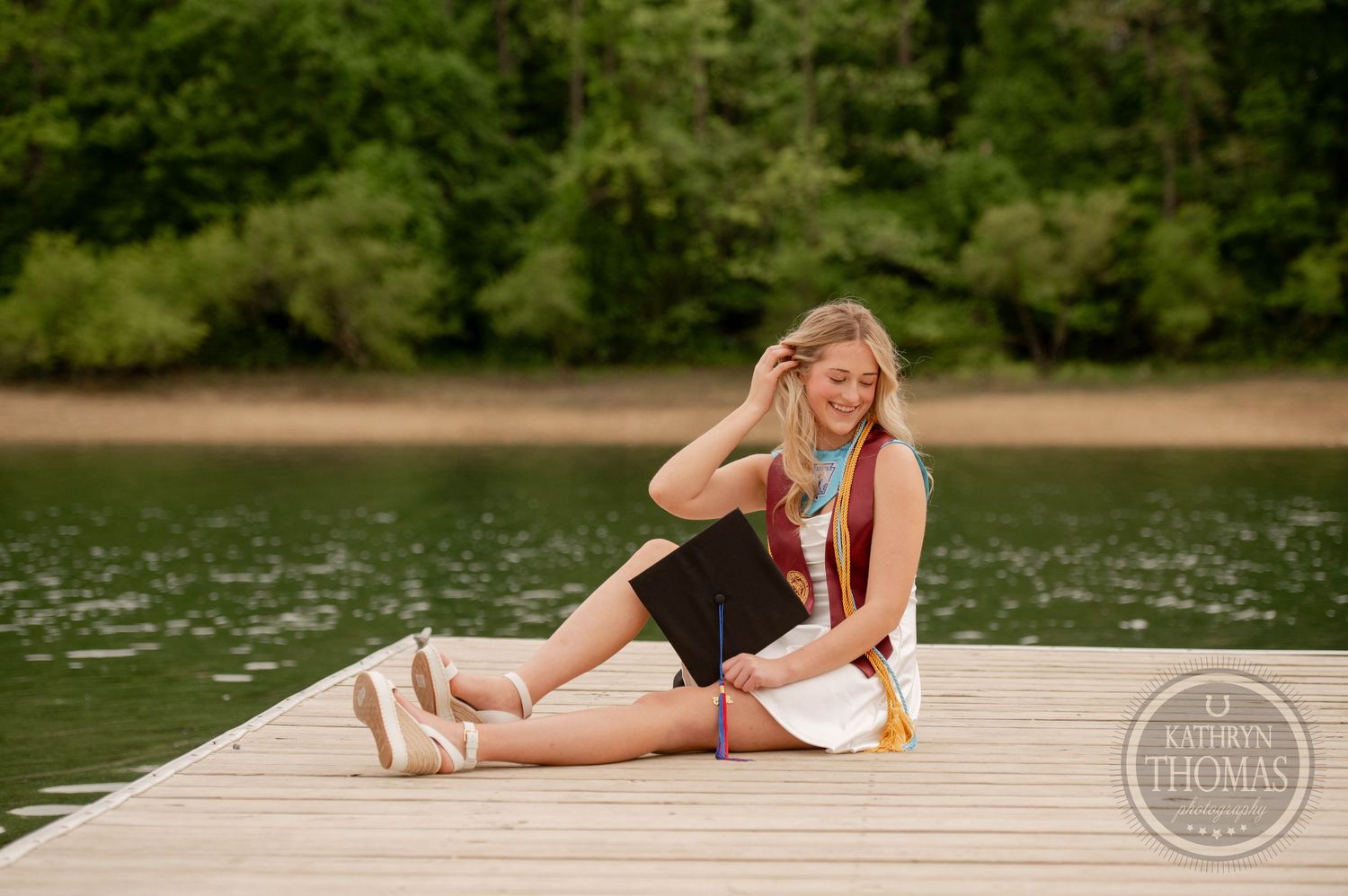 A person in black attire sits on a wooden dock by a lake during summer with trees in the background.