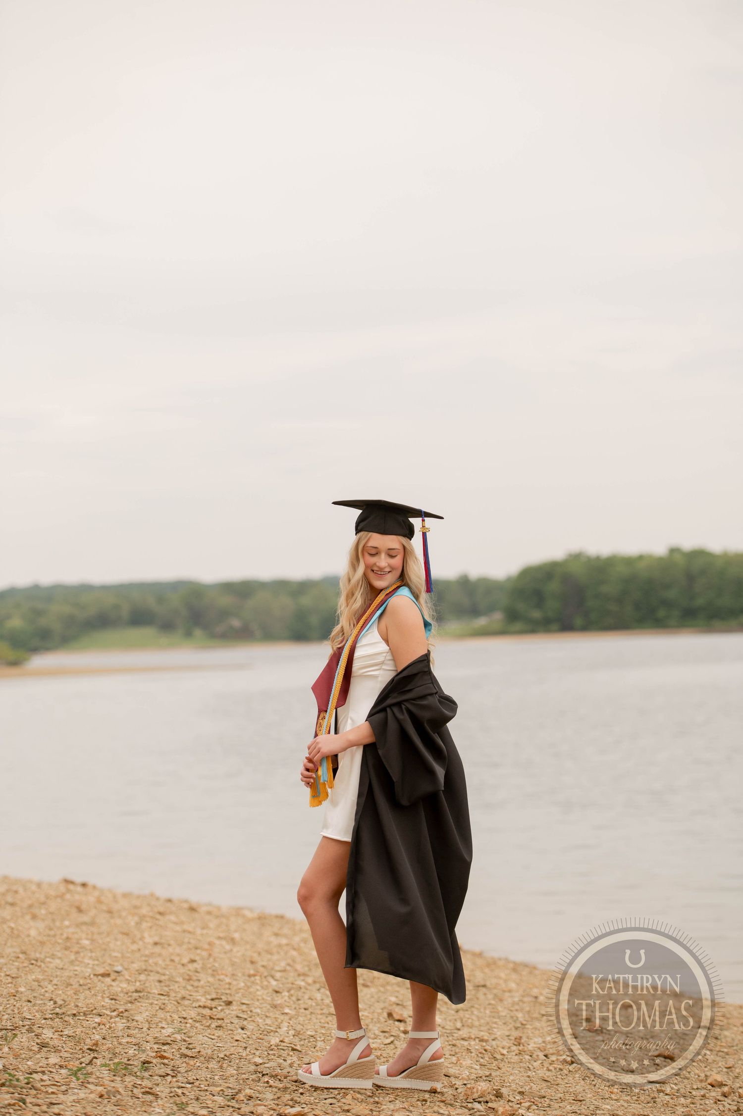 Graduate in black cap and gown poses for portrait on sandy beach shore with lake and trees in background.