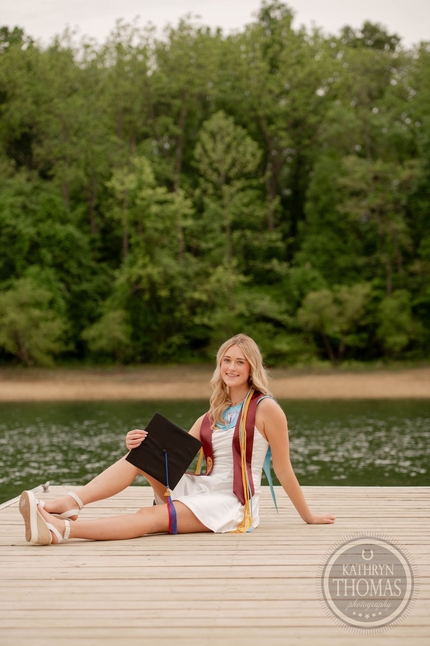 Graduate in white dress and brown vest sits on wooden dock by lake surrounded by green trees on sunny day.