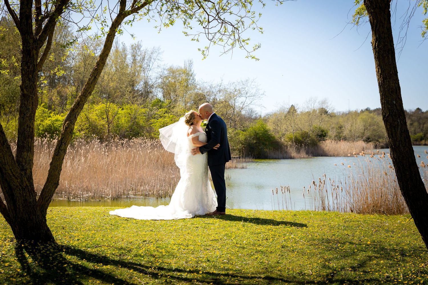 Bride and groom share a kiss by the lake at The Village Hotel, surrounded by nature and spring light.