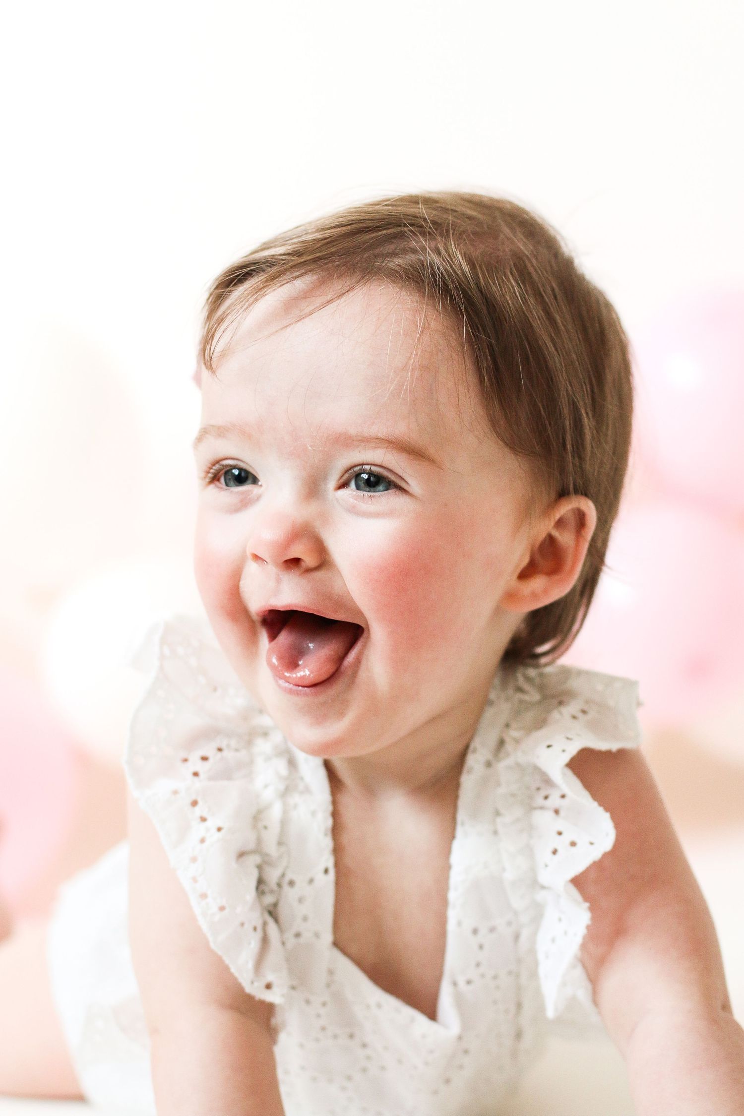 A joyful baby wearing a white eyelet lace outfit smiles brightly against a pink background, during birthday shoot in Nottingham