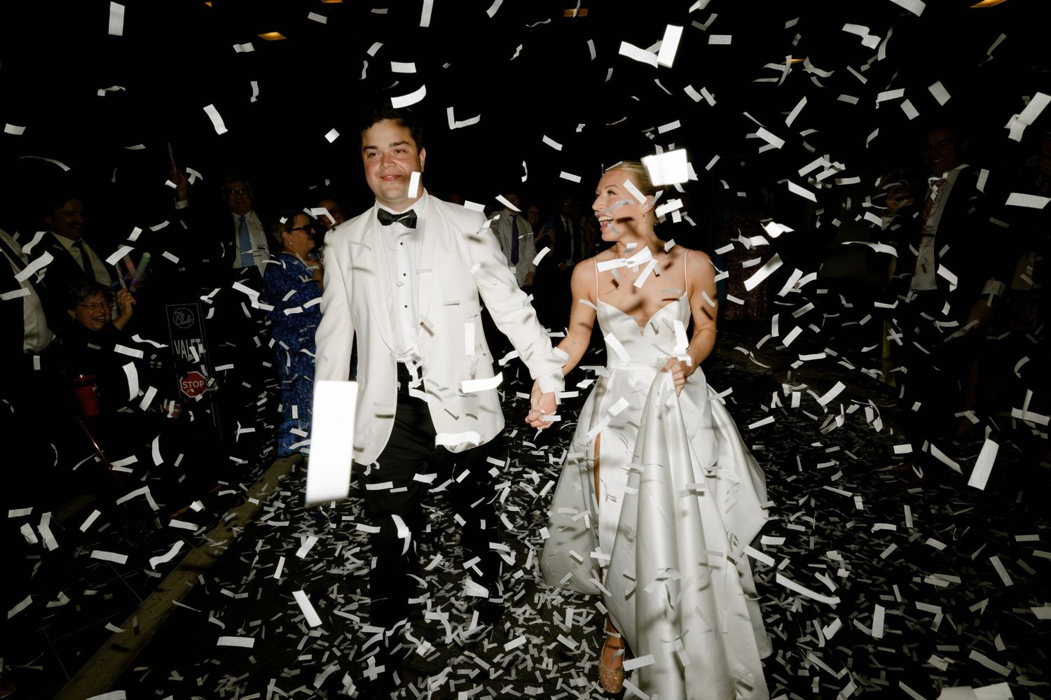 A wedding couple in white formal attire celebrate amid falling confetti during a festive reception entrance.