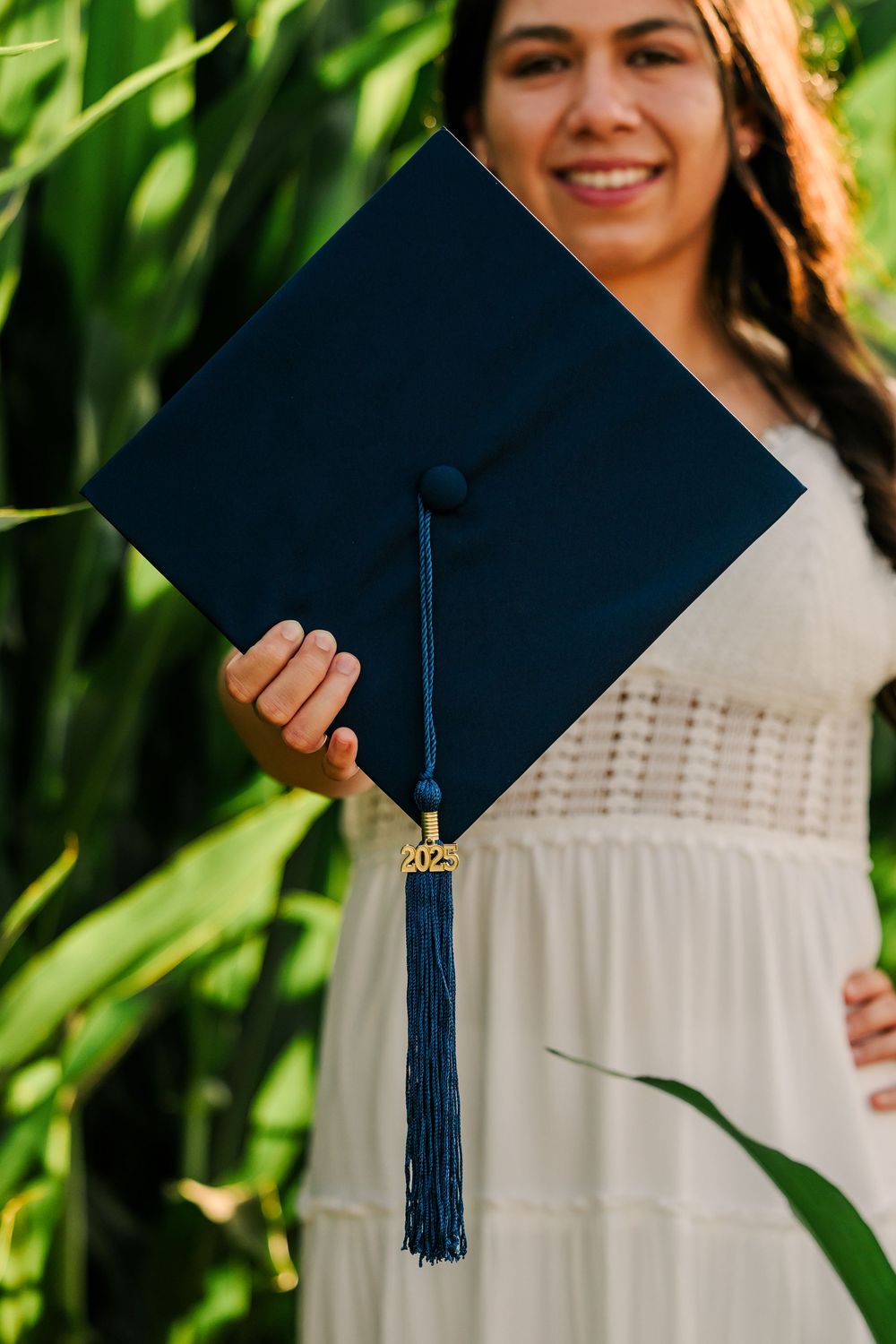 Graduate holds up navy blue graduation cap with tassel against lush green foliage in white dress outdoors.