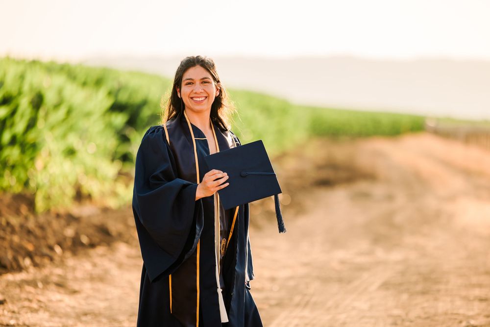 A graduate in academic regalia smiles while holding their diploma on a sunny outdoor path.