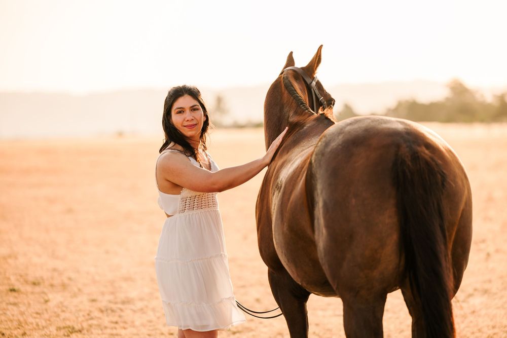 Someone in a white dress stands beside a brown horse in a dusty field at sunset.