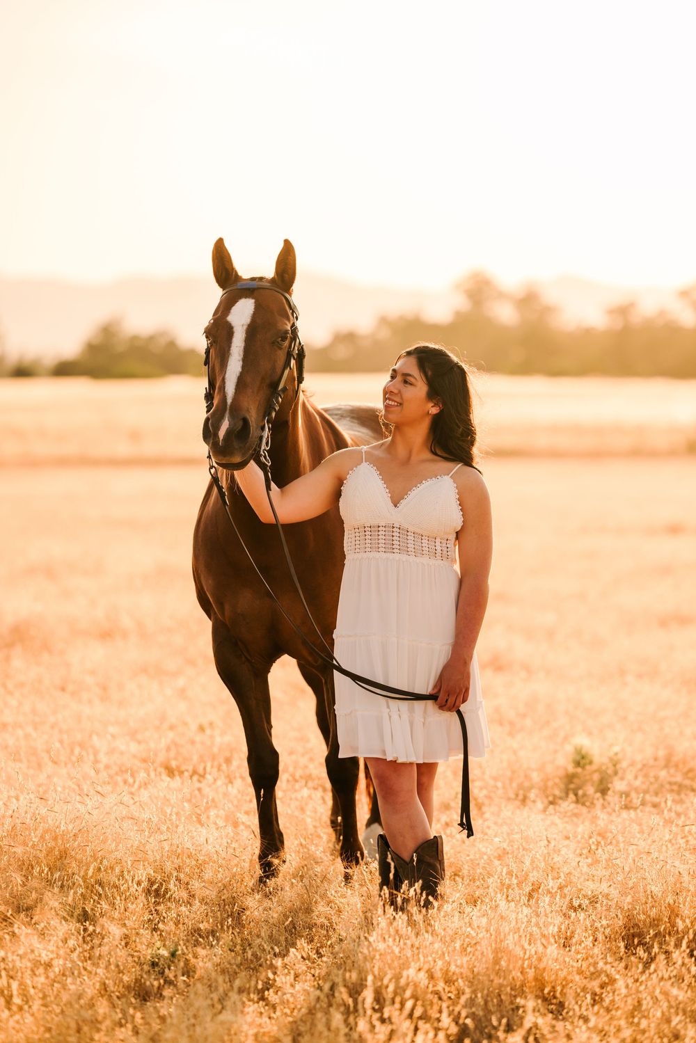 A woman in white dress stands beside her bay horse in a sunlit meadow during golden hour.
