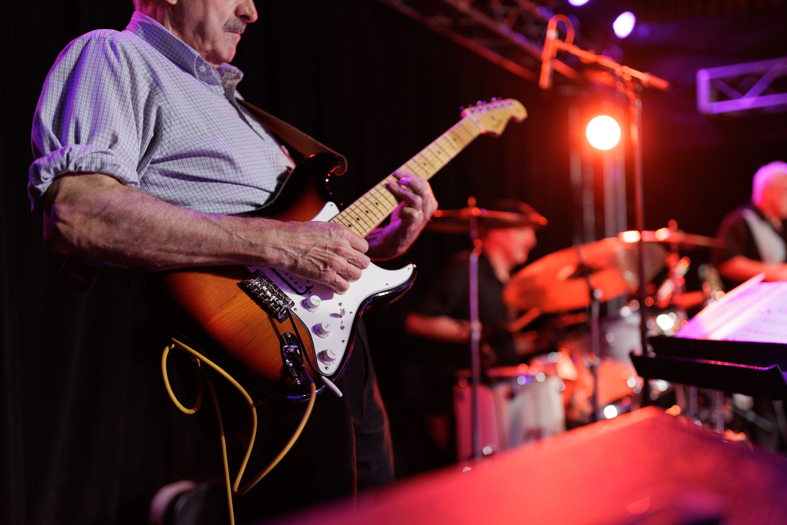 Musician playing electric guitar on stage with red lighting and drum kit visible in background.