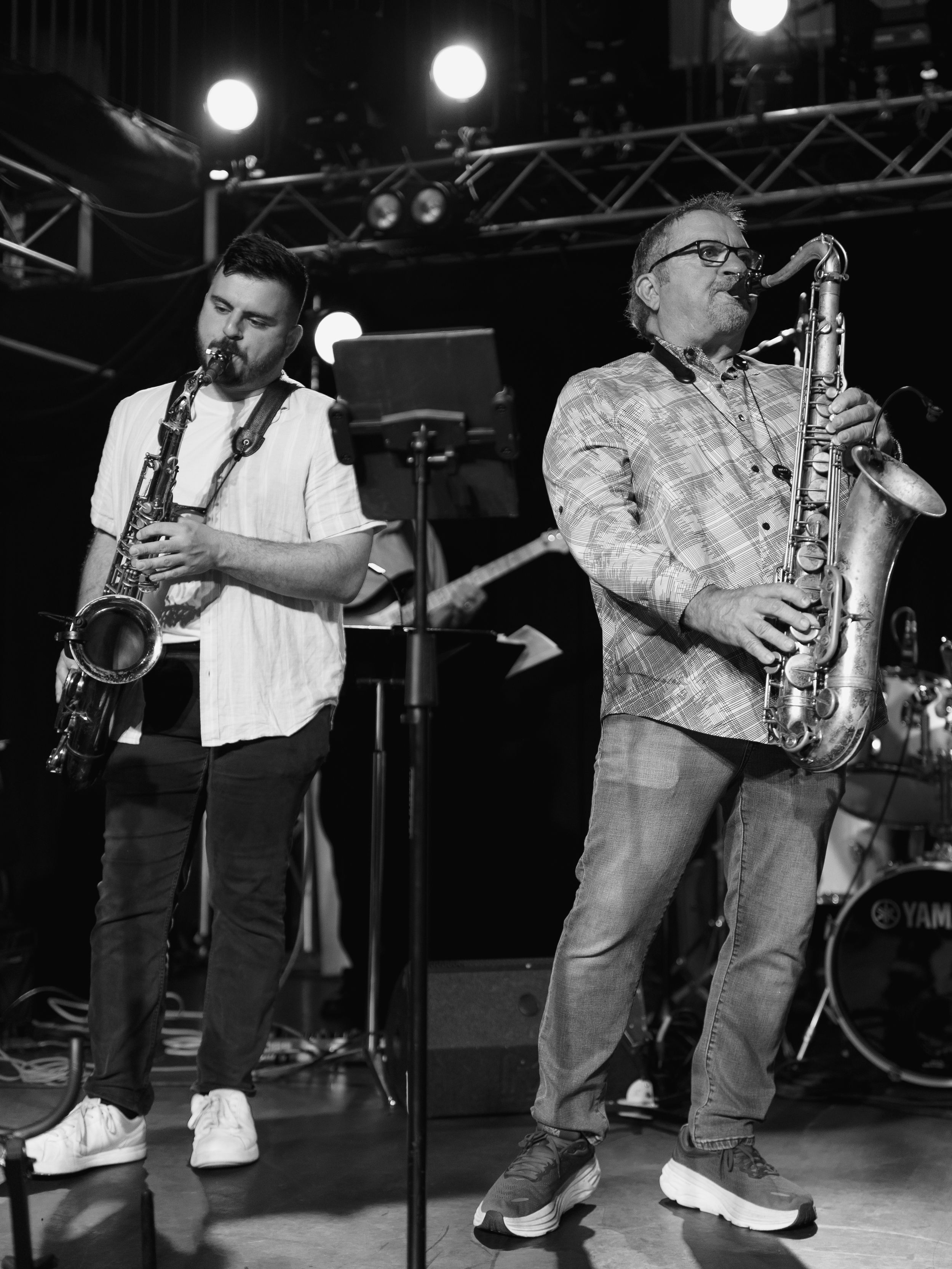 Two saxophone players perform together on stage under bright spotlights during a jazz performance.