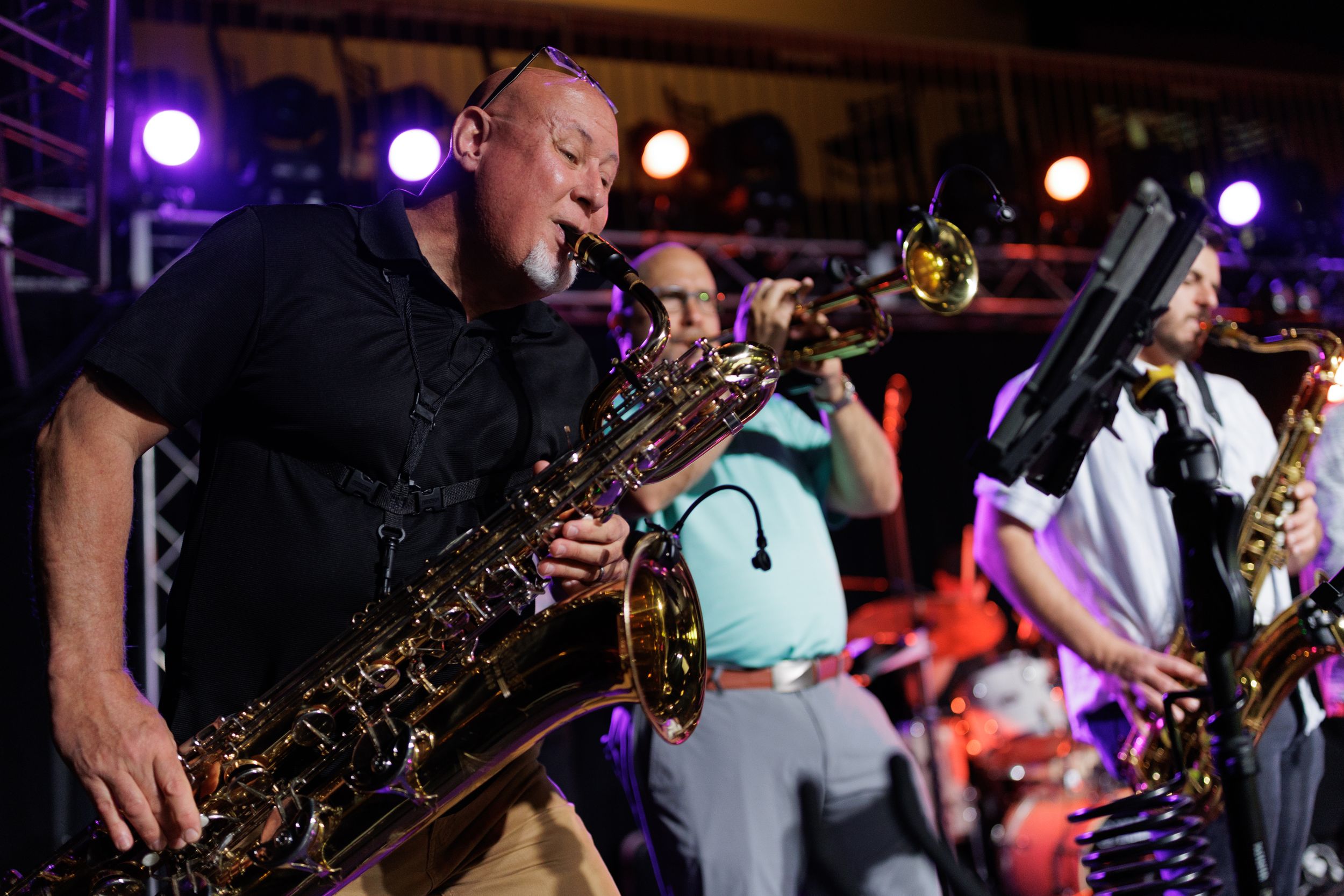 A saxophonist performs on stage with a band under purple stage lighting during a live musical performance.