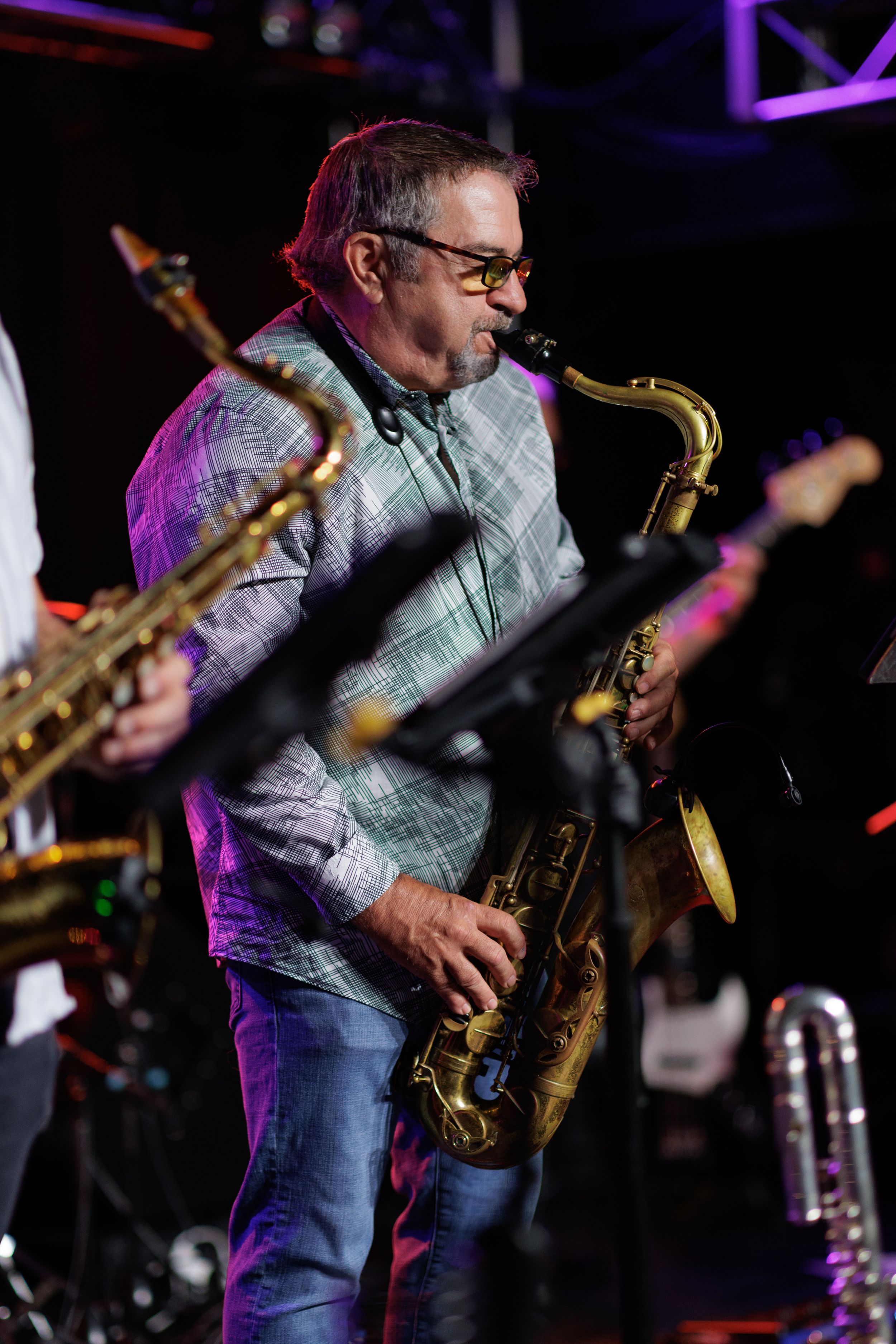 Saxophone player wearing glasses and gray shirt performs passionately on stage under purple lighting.