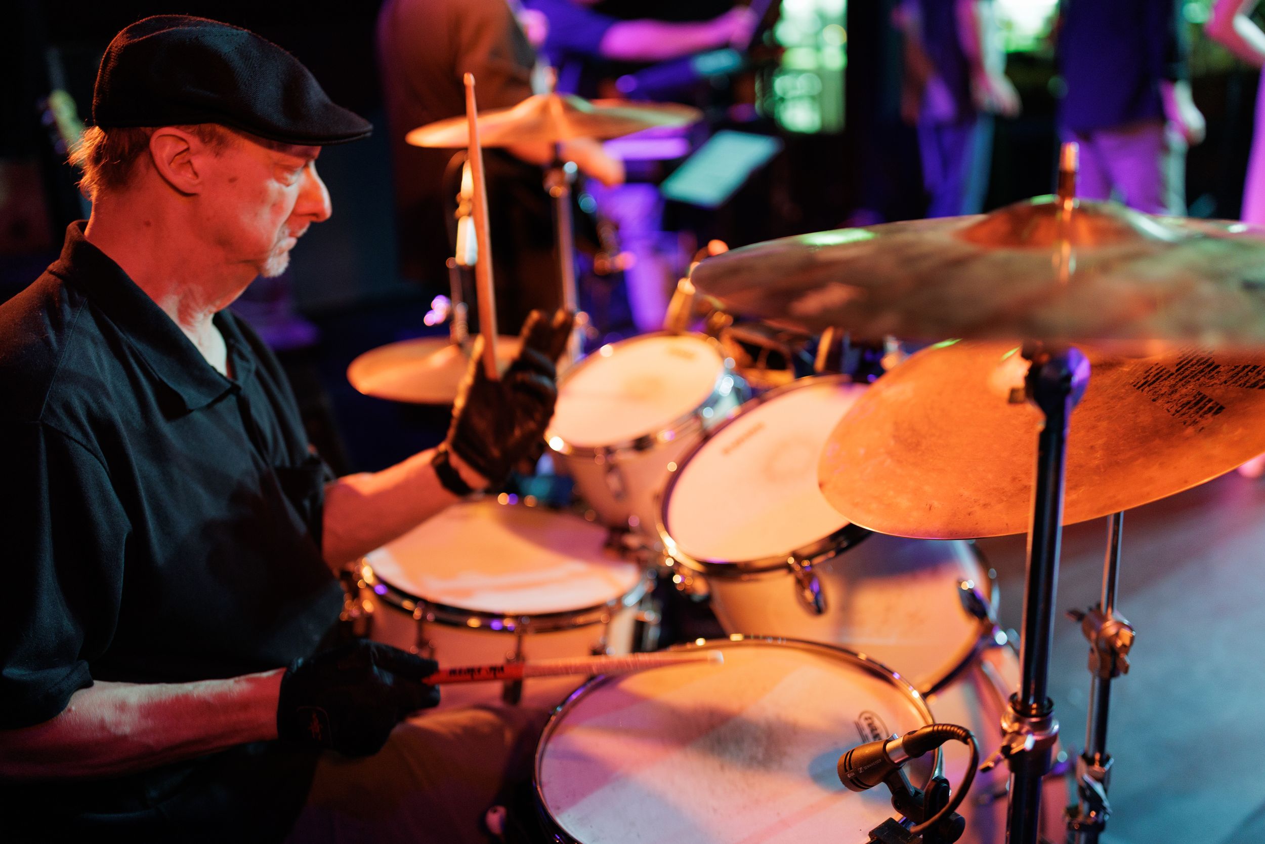 Drummer in black cap passionately plays drumset on dimly lit stage with colorful lighting in background.