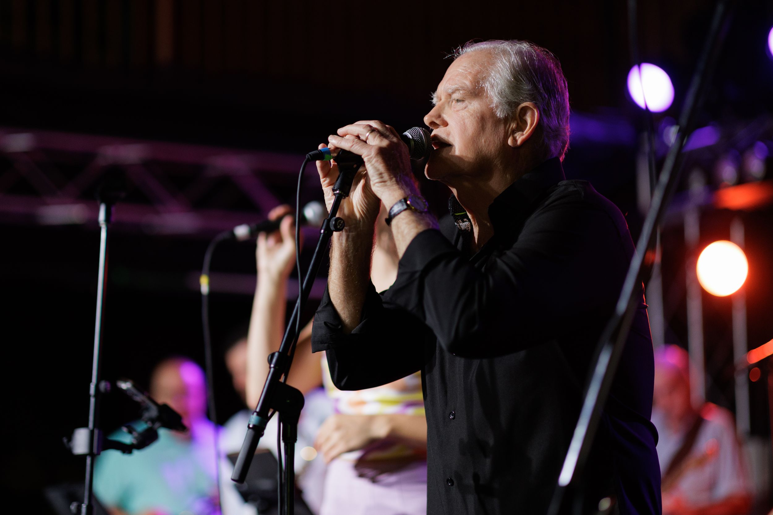 A performer in black attire sings passionately into a microphone during a live concert with purple stage lighting.