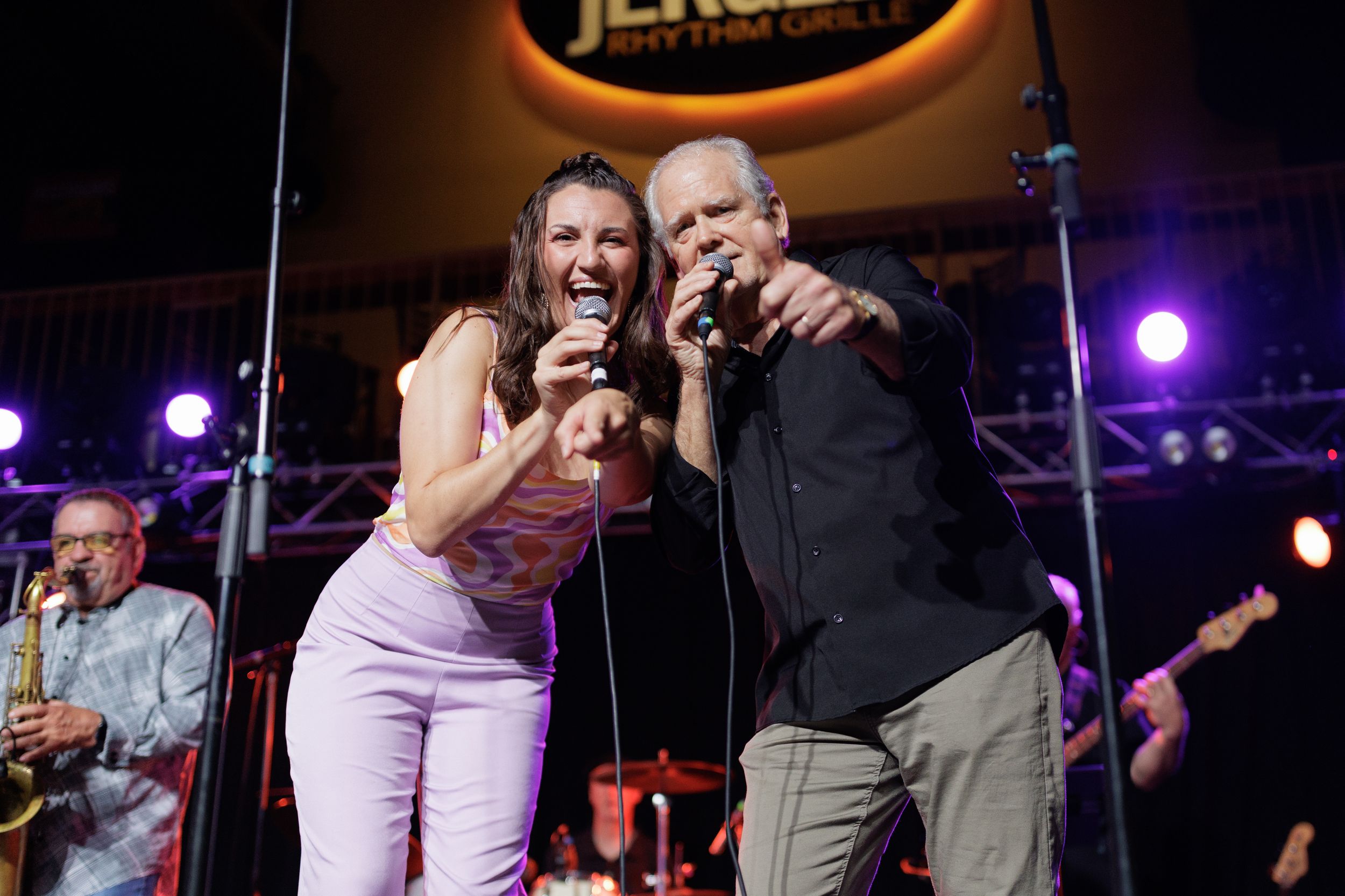 Two performers share a microphone on stage during an energetic nightclub performance with purple lighting.