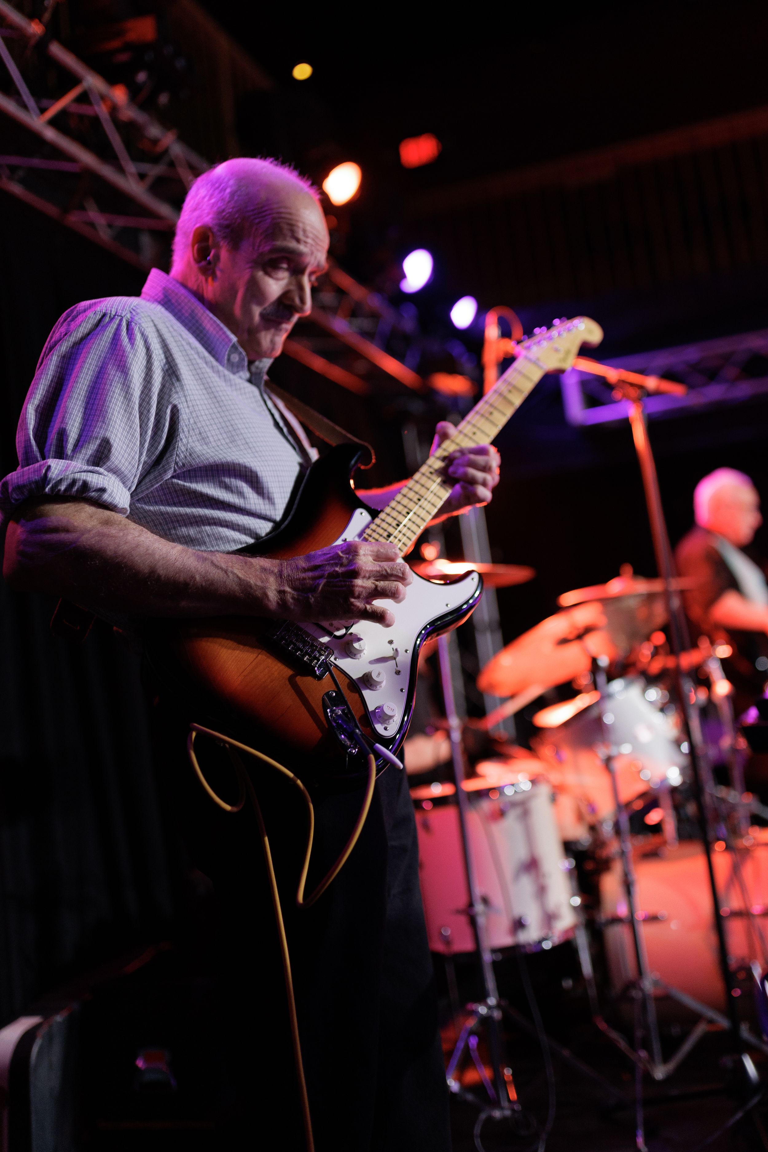 Guitarist playing electric guitar on dimly lit stage with purple and red stage lighting effects.