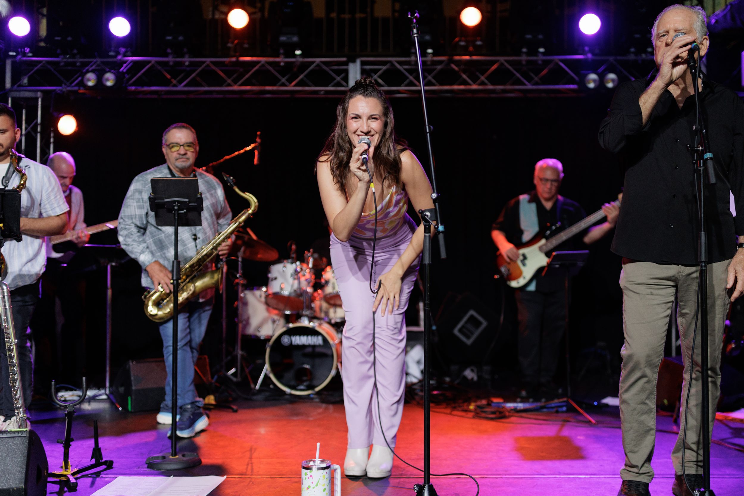 Musicians perform on stage under purple and red lighting during an outdoor concert performance.