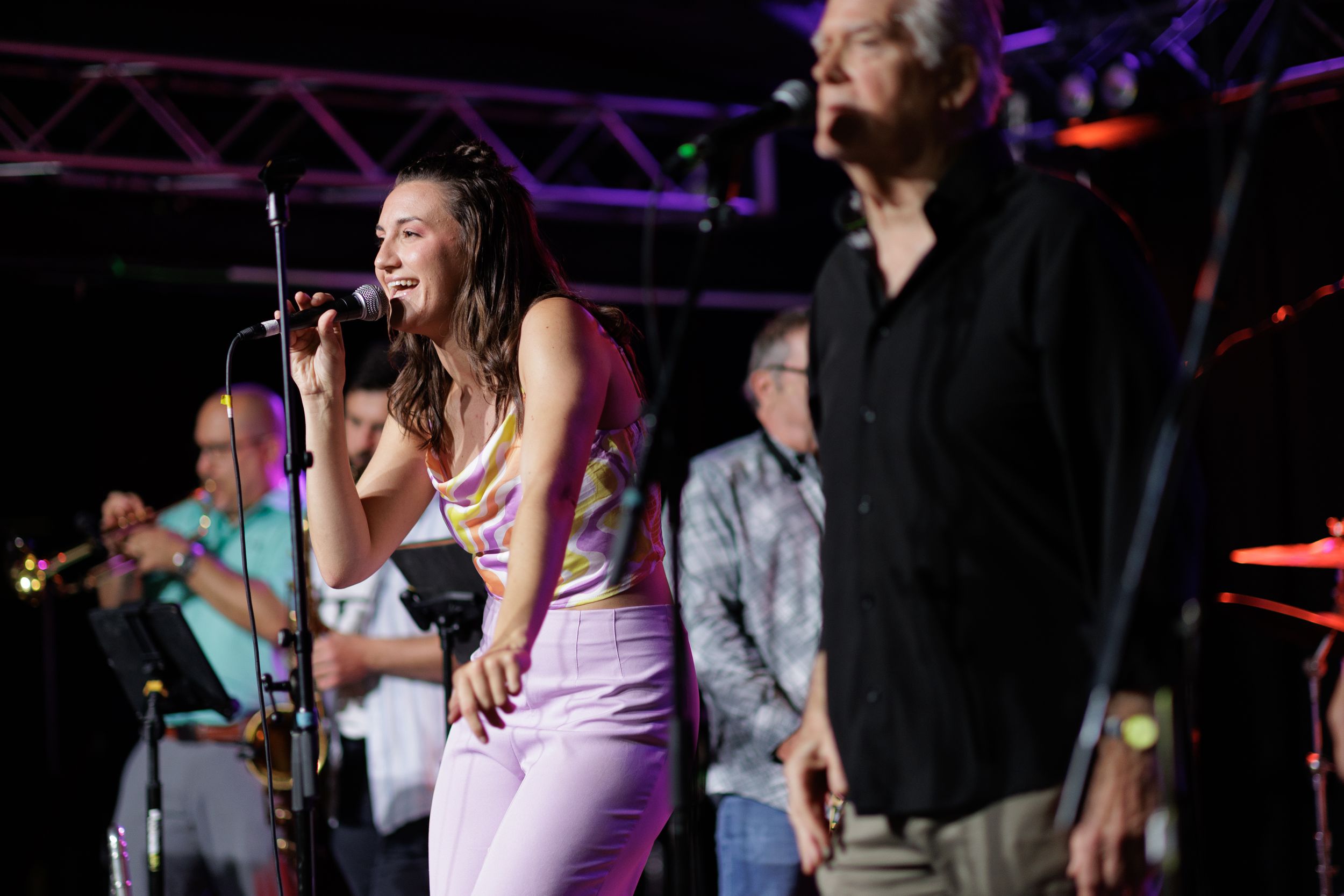 Performers on stage at a live music concert with purple lighting and sound equipment visible in the background.