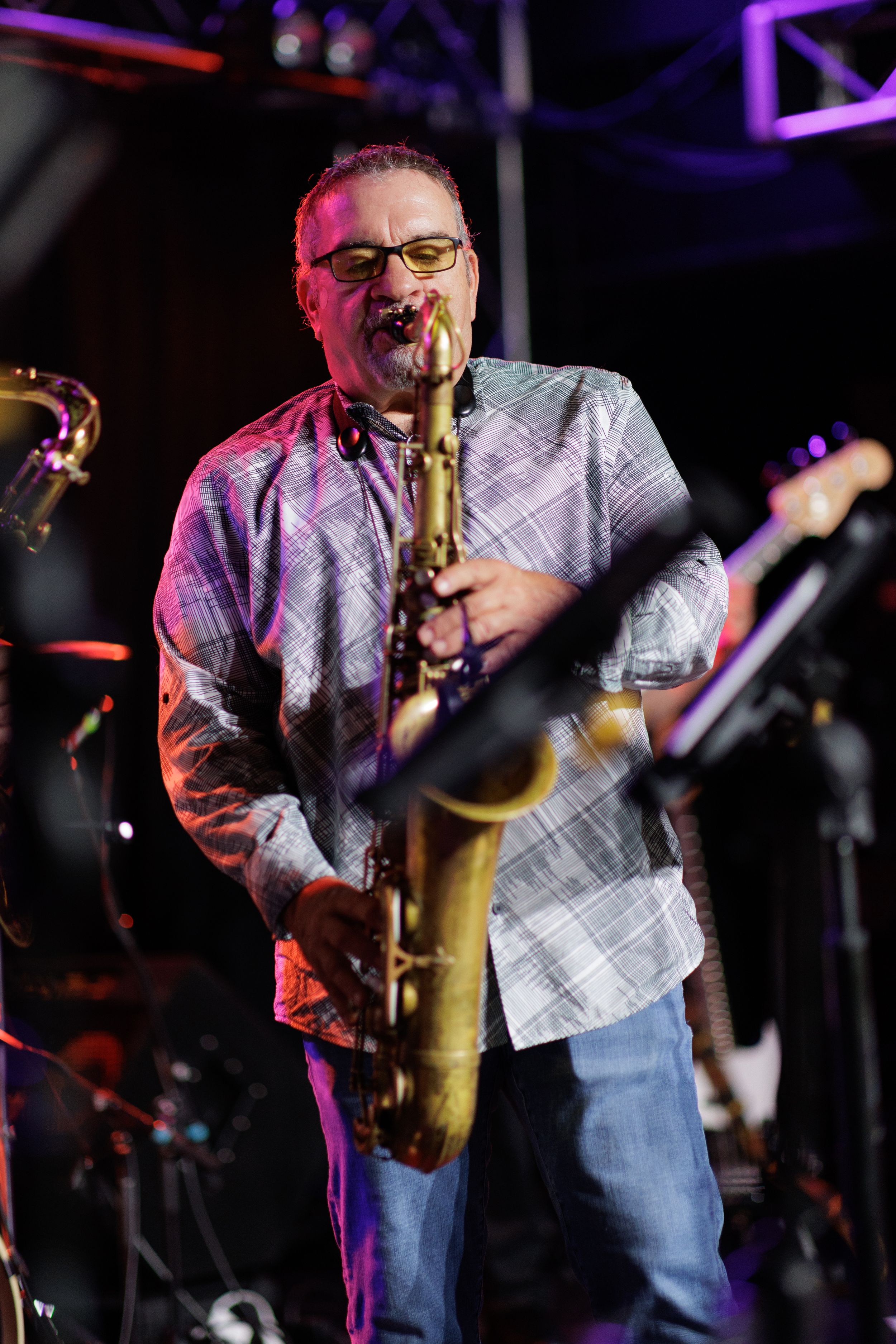 A saxophone player performs on stage illuminated by purple and red stage lighting during a concert.