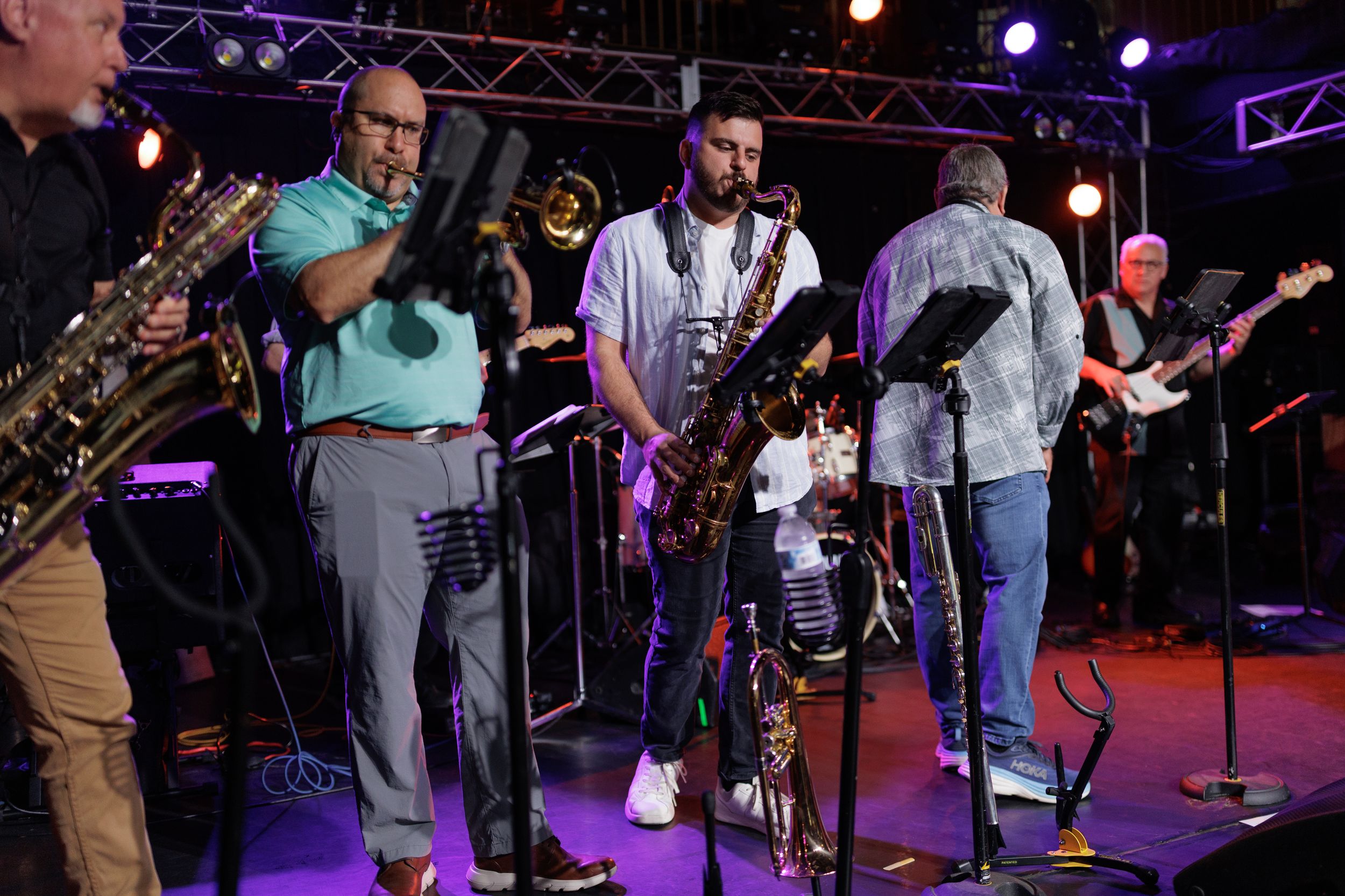 Musicians perform on stage with saxophones, guitars and other instruments under colorful stage lighting at a live concert.