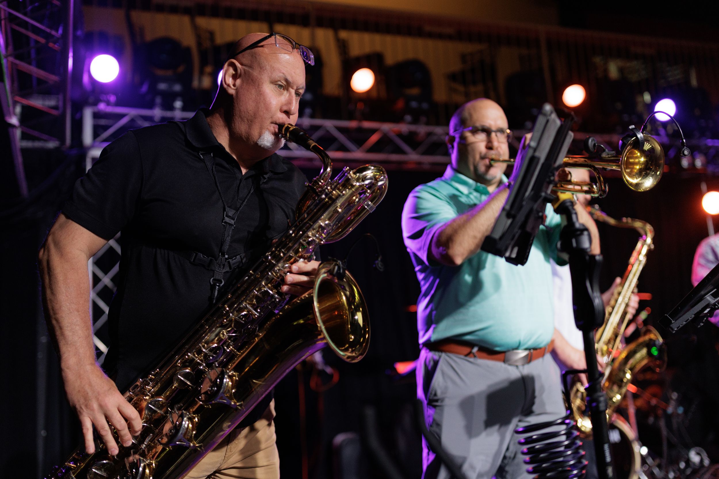 Musicians perform on stage with brass instruments under colorful purple and red stage lighting during a live concert.