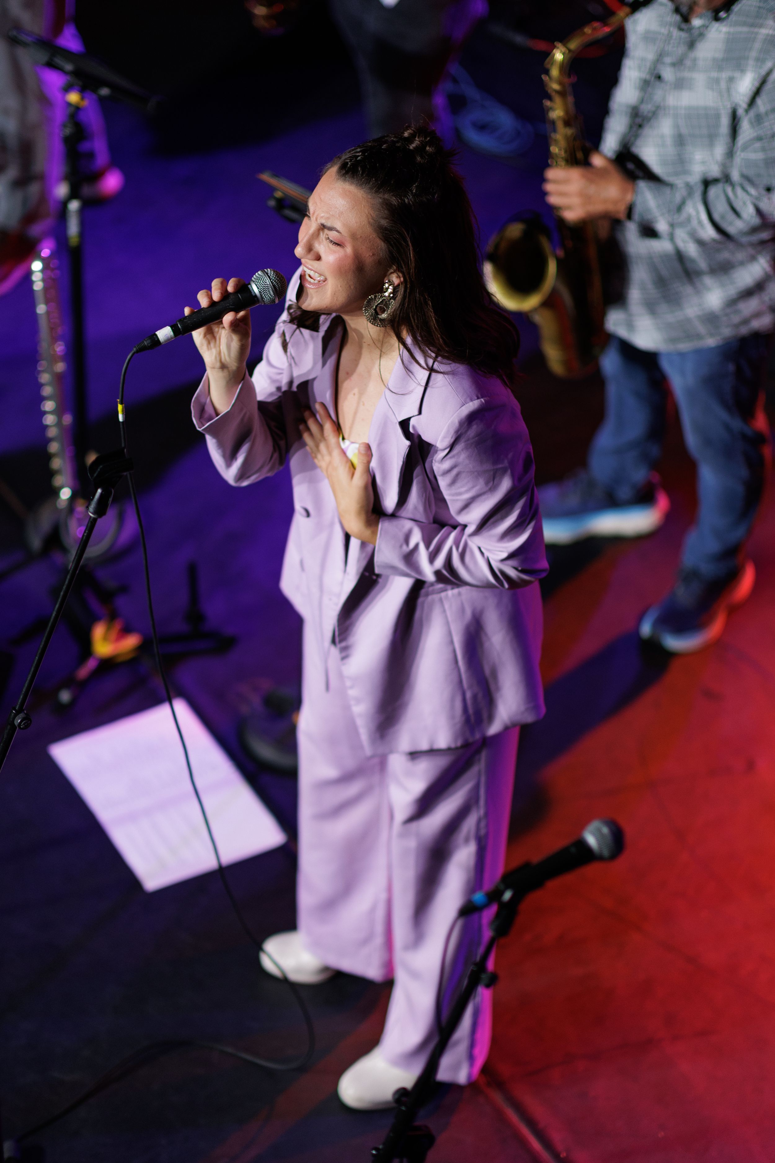 A performer in a purple suit sings passionately into a microphone on stage with band members visible in the background.