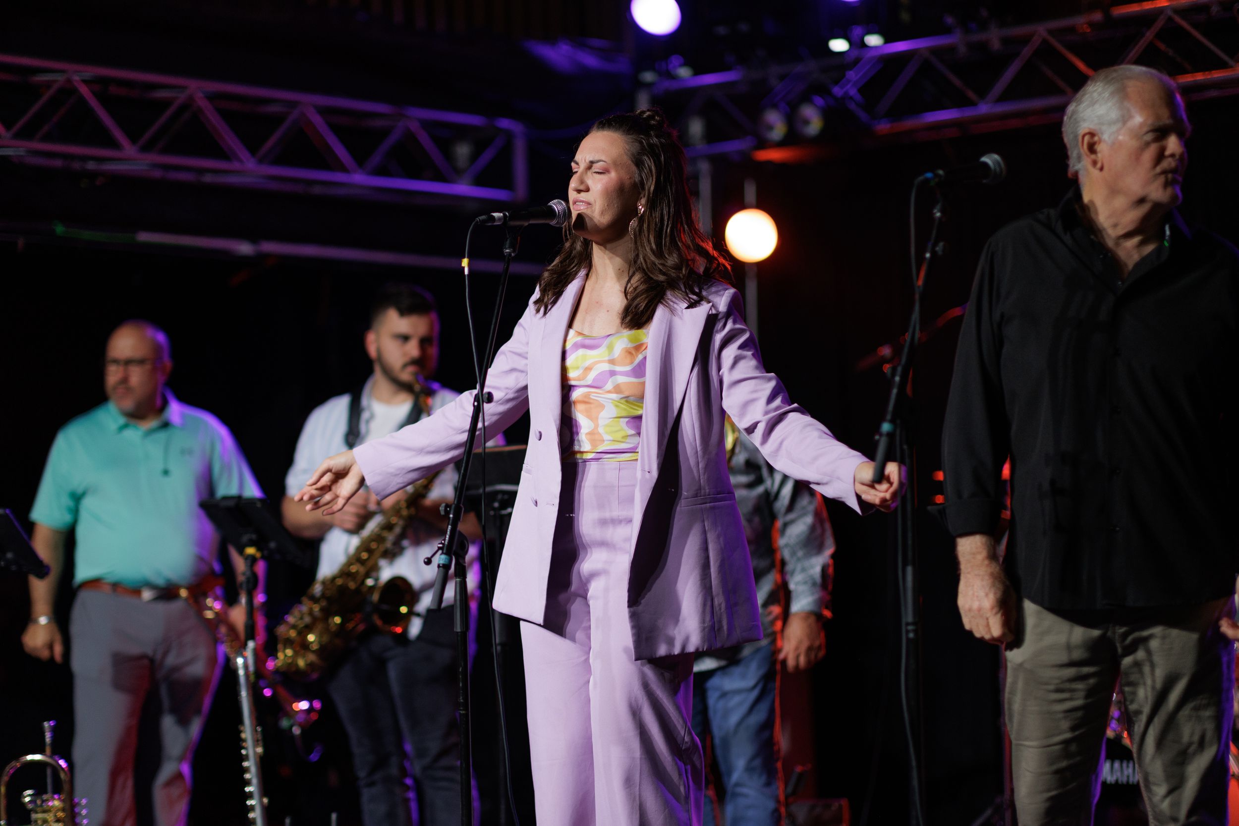 A performer in a purple outfit sings on stage with band members and lighting equipment visible in the background.
