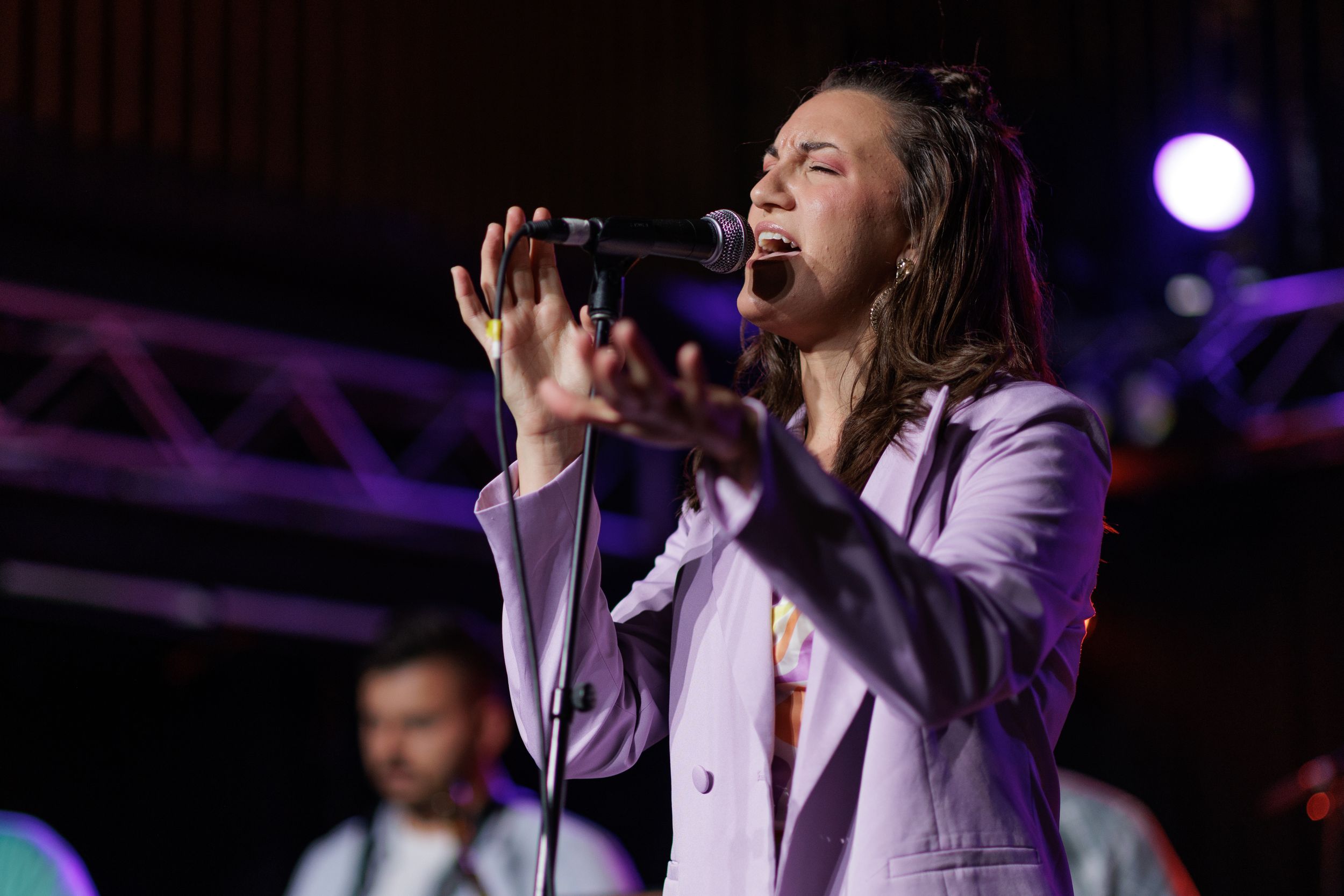 A performer in a light-colored suit sings passionately on stage with dramatic lighting effects during a live concert performance.