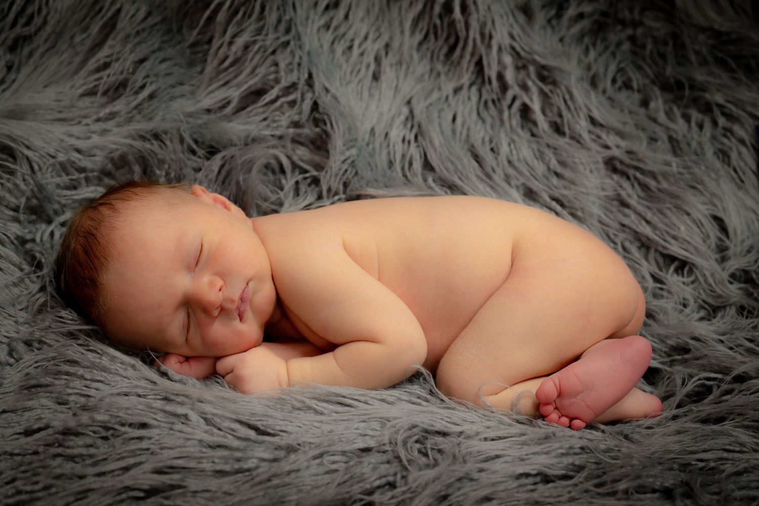 Peaceful sleeping newborn captured in artistic nude portrait on gray textured fur background.