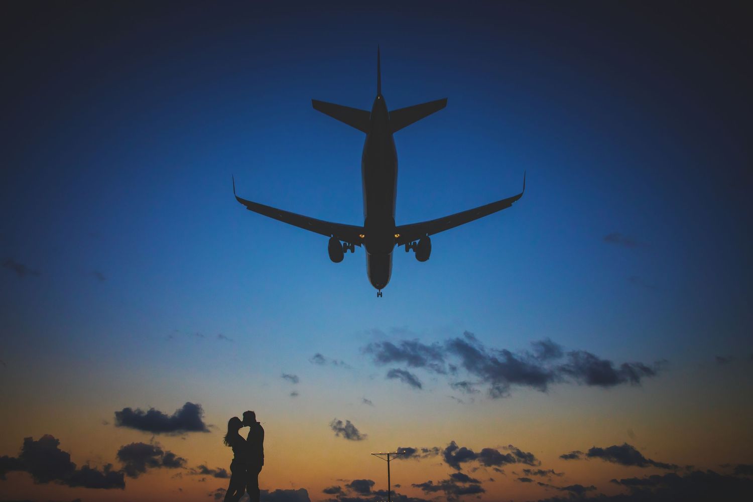 A silhouetted passenger airplane flies overhead against a dramatic sunset sky while a couple embraces below.