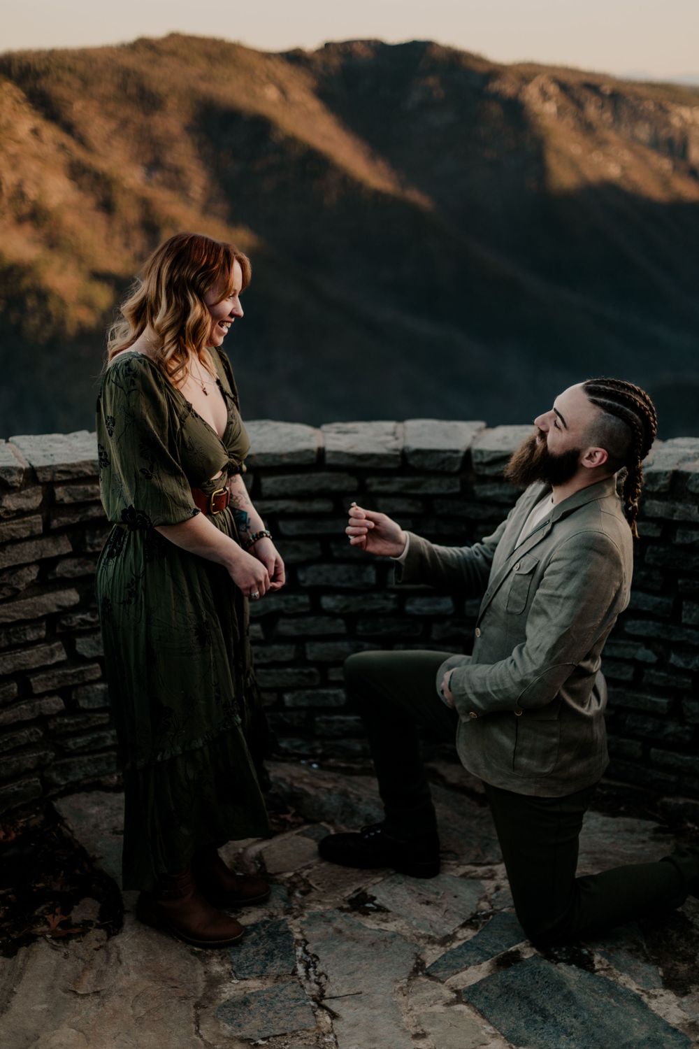 Romantic proposal scene at sunset with mountains and stone wall in background.