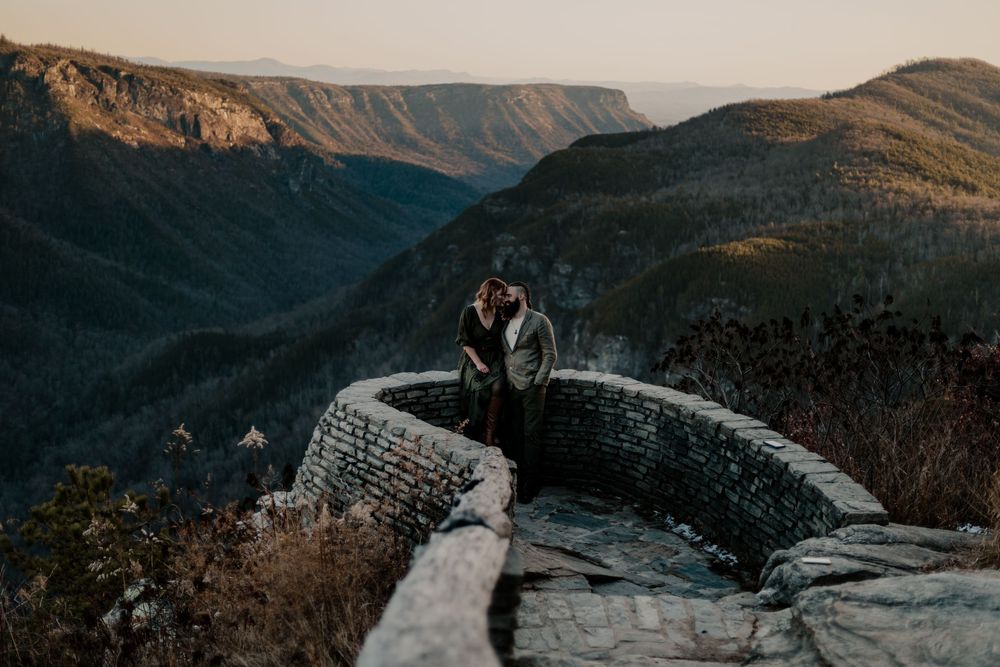 Couple embracing on mountain overlook during golden hour with dramatic landscape views.
